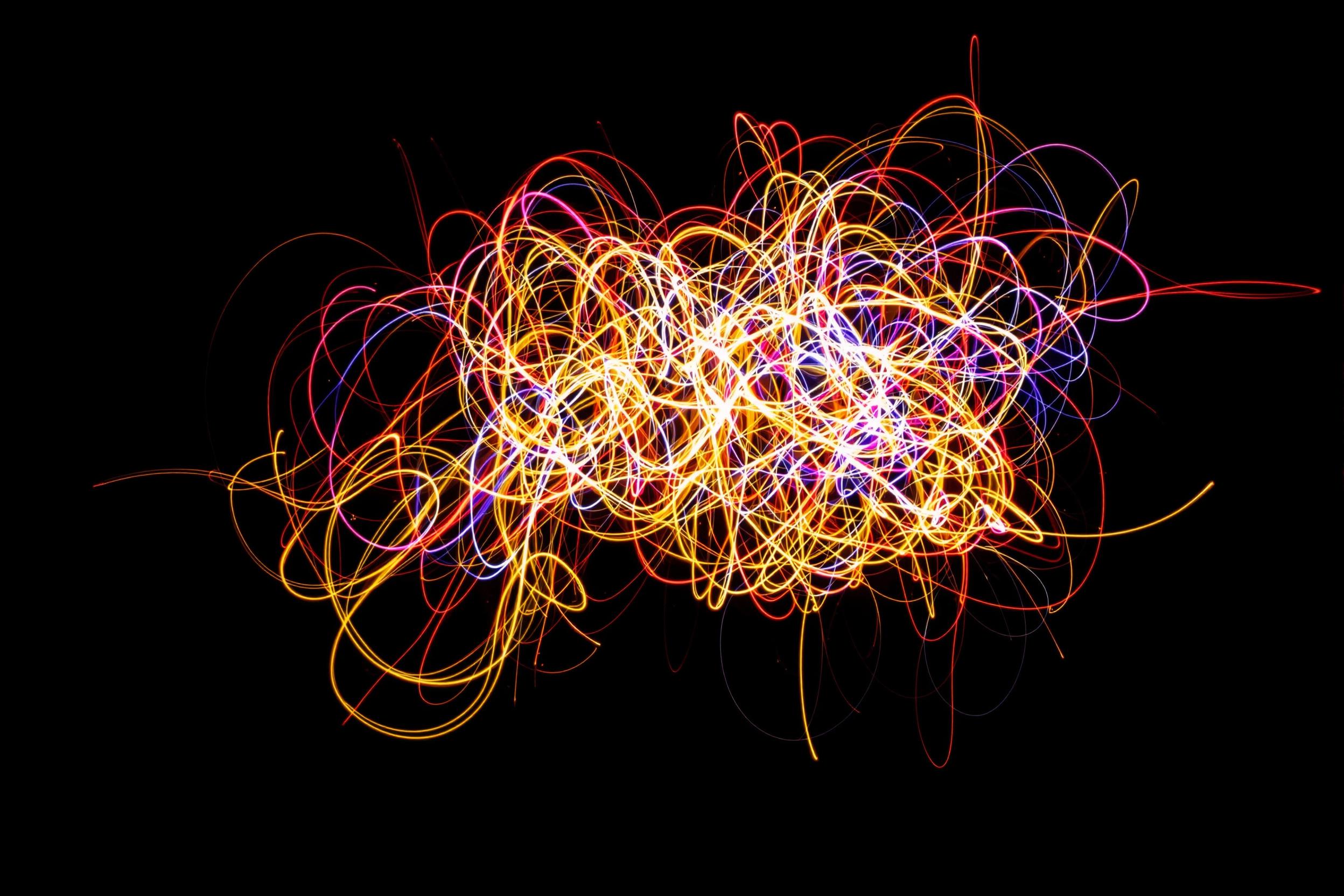 An abstract light painting bursts with energy, captured through a long exposure. Trails of multicolored light form intricate loops and patterns against a black background. Bright tones of red, yellow, blue, and purple contrast sharply, creating an electrifying spectacle. Subtle motion creates a sense of dimensionality, giving depth to the swirling forms.