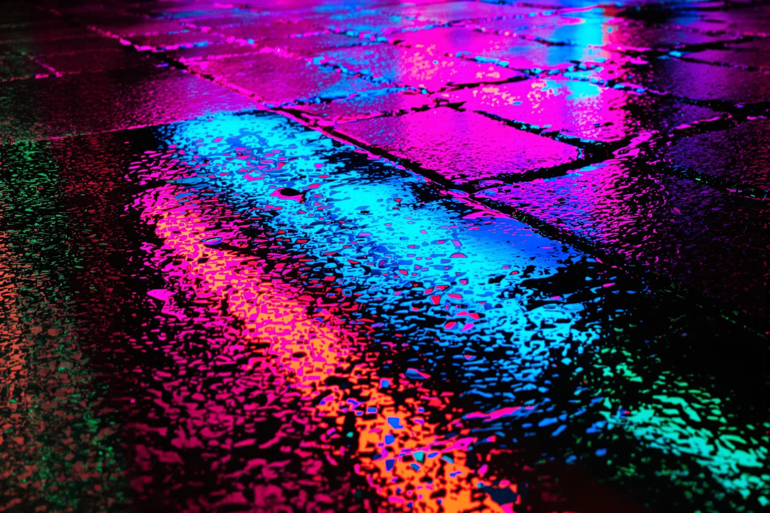 A close-up of a rain-slicked urban sidewalk at night reflects vibrant neon lights in bold shades of pink, blue, and green. The abstract composition features glistening textures and diagonal streaks of water. Puddles distort the sharpness of reflected signage, adding a dynamic play of color and shimmer.