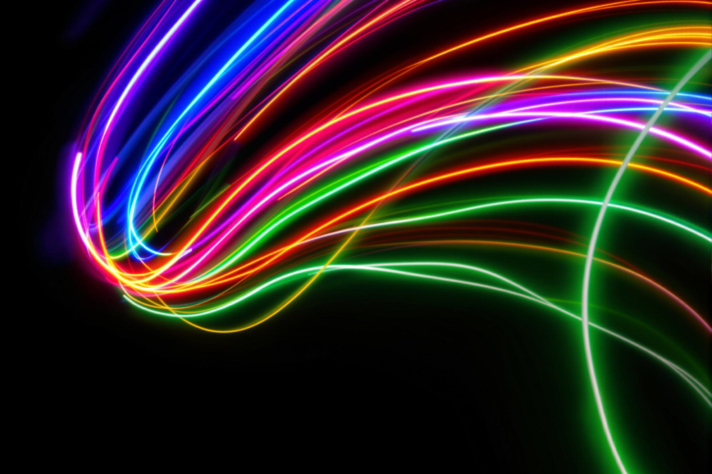 Streaks of multicolored neon lights arc dynamically across a pitch-black background in layered, curving trails. The vivid hues of blue, pink, orange, and green form an abstract, flowing composition captured with deliberate motion blur. Contrast is sharp, evoking a sense of energy and boundless creativity.