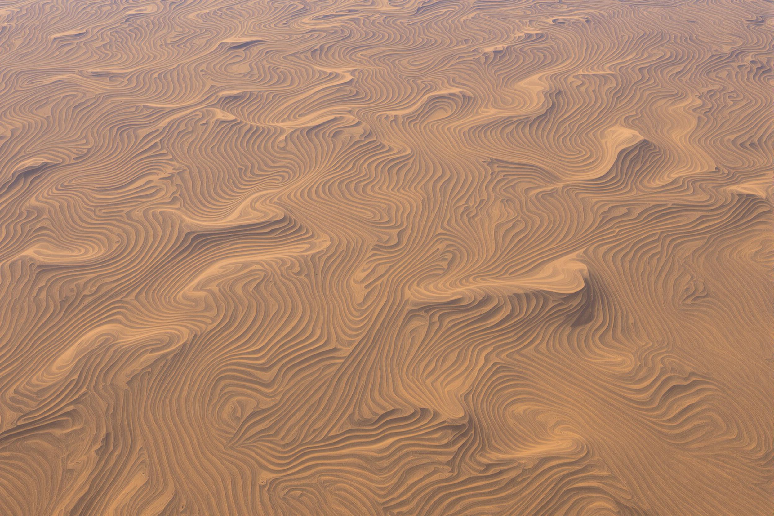 Discover the mesmerizing beauty of wind-sculpted sand dunes in this abstract aerial photograph. Sinuous curves and ripples create a hypnotic landscape, showcasing nature's artistry in earth tones.