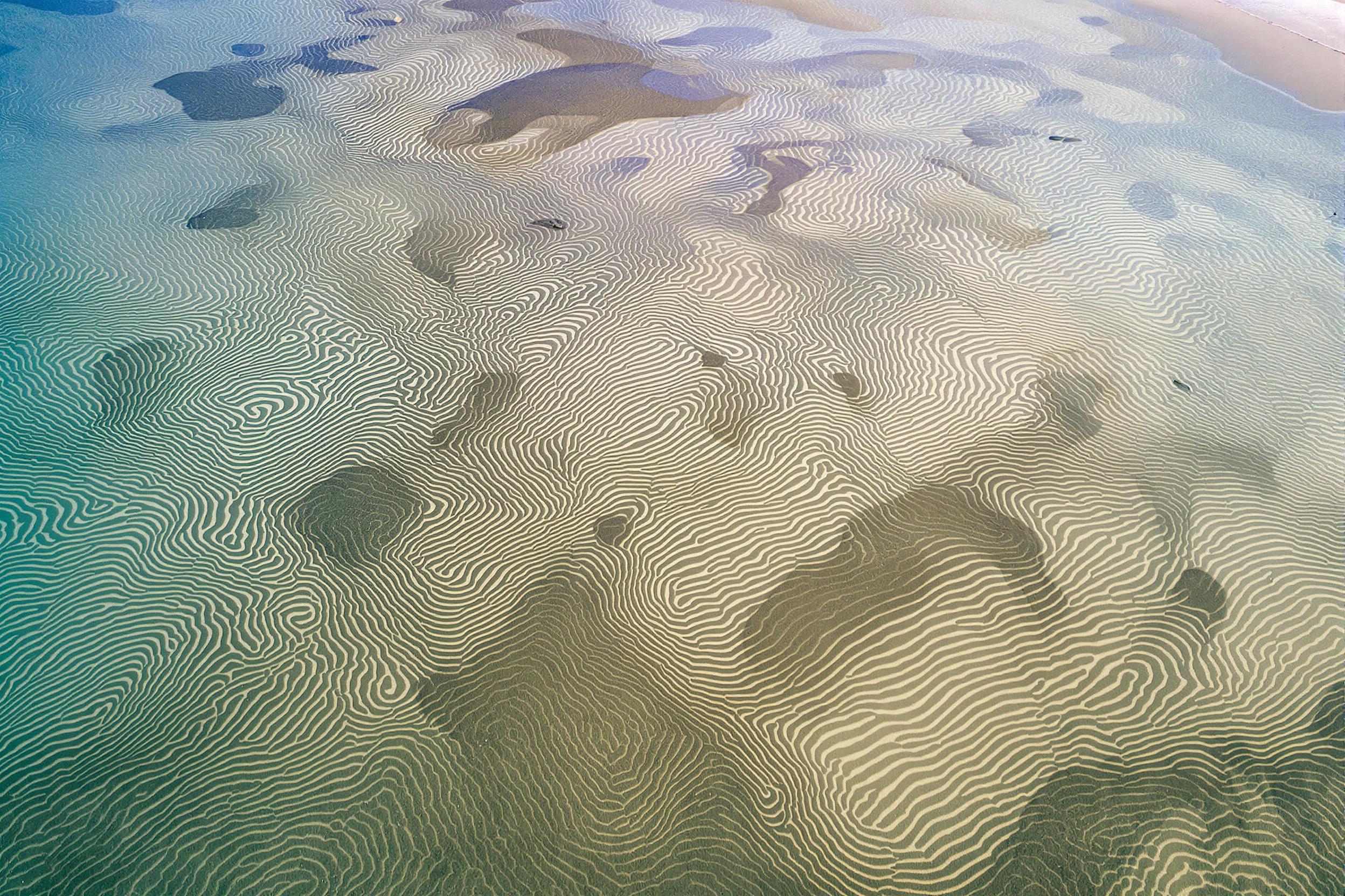 An aerial view unveils mesmerizing tidal patterns etched into a sandy coastal sandbar. Delicate ripple textures interlace with shallow pools of water, reflecting a gradient blue sky. Soft shadows delineate swirling formation edges under even midday sunlight, showcasing nature’s artistry in organic symmetry.