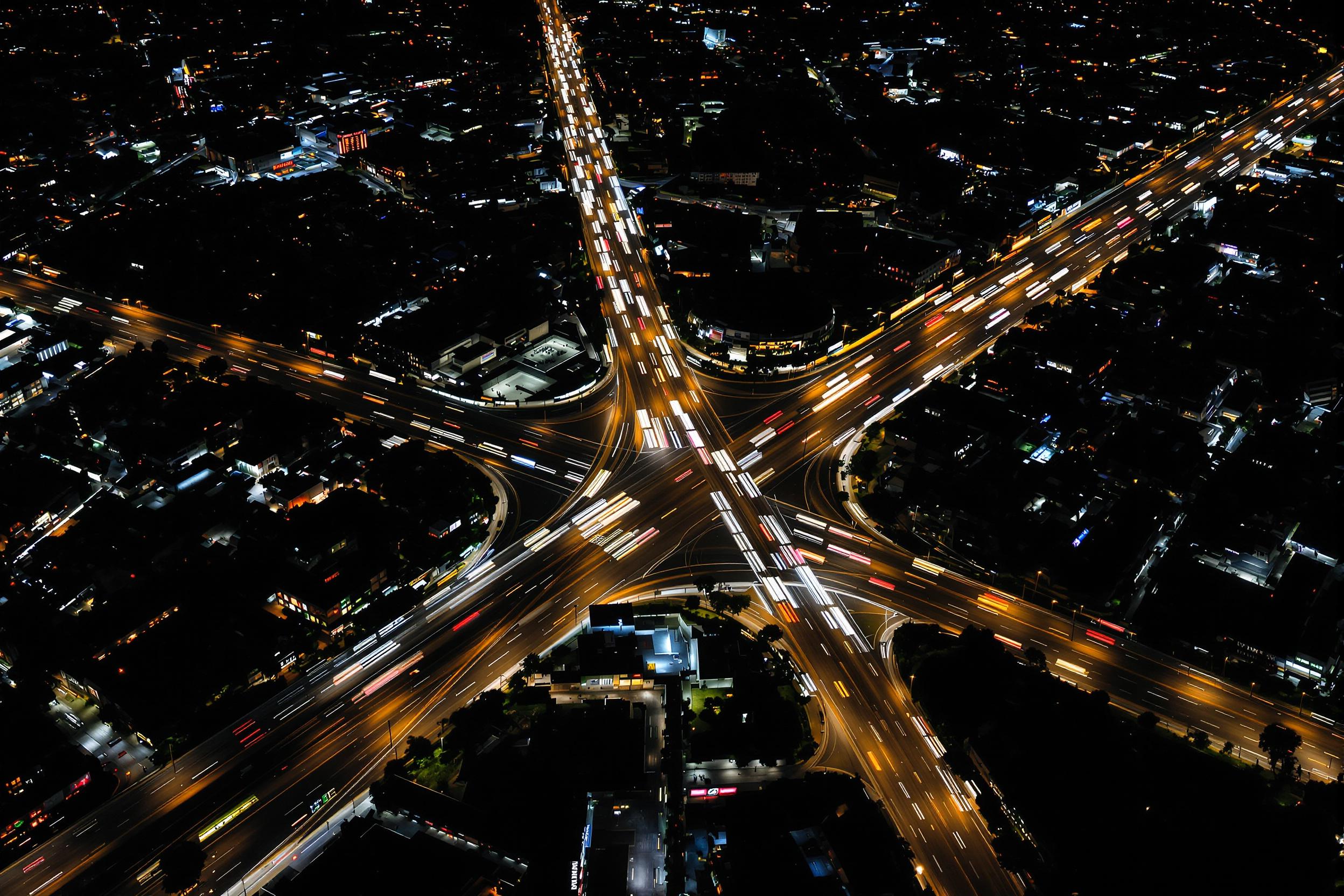 Stunning nighttime aerial photograph of a bustling city intersection. Long exposure captures the flow of traffic as streaks of light, creating a dynamic web against the dark urban landscape. Perfect for showcasing modern city life or transportation themes.