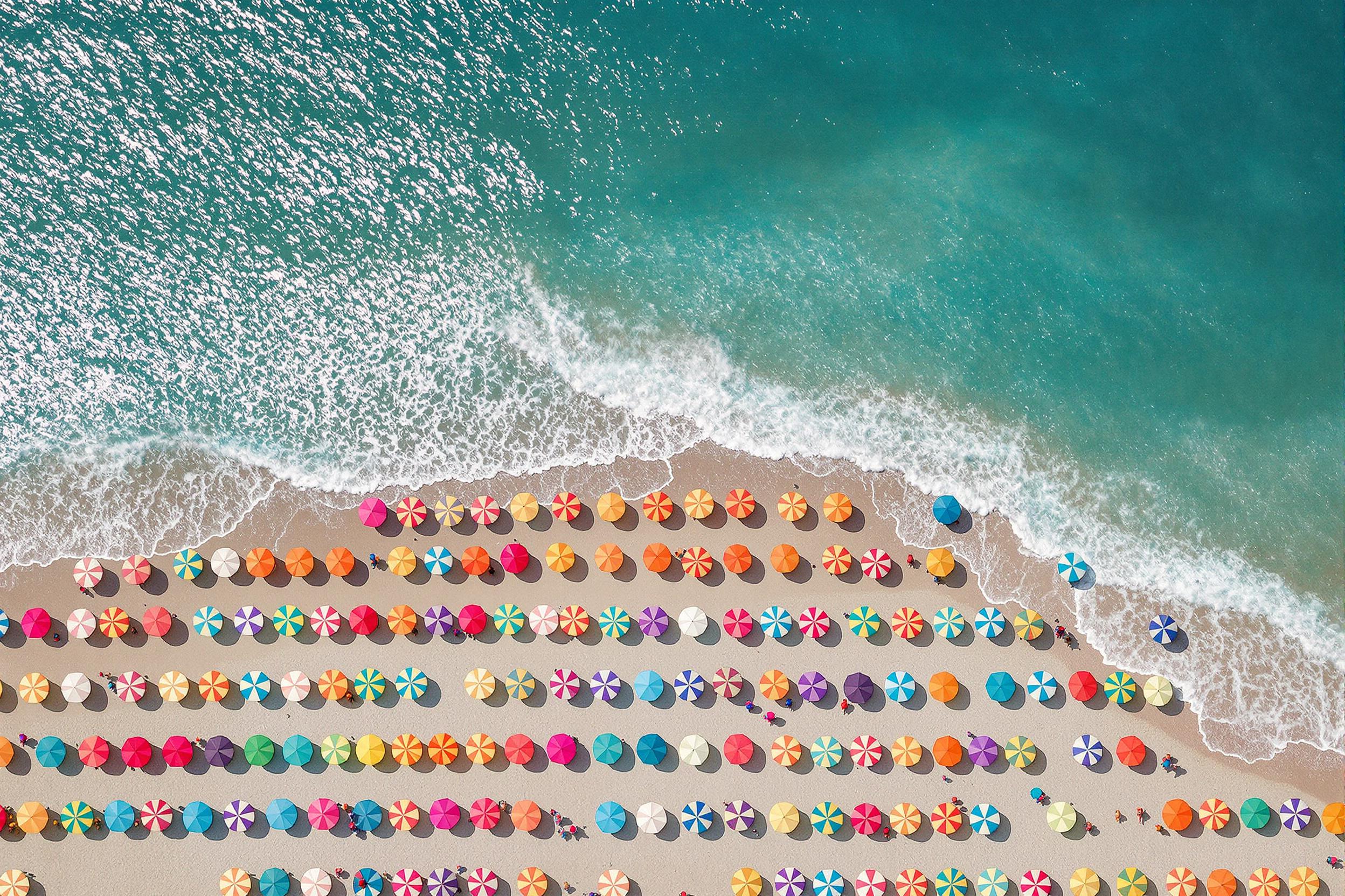 Vibrant aerial image capturing a sea of multicolored beach umbrellas dotting a sandy shoreline. The geometric patterns formed by the umbrellas create a mesmerizing visual against the azure waters. Ideal for summer vacation or coastal lifestyle concepts.