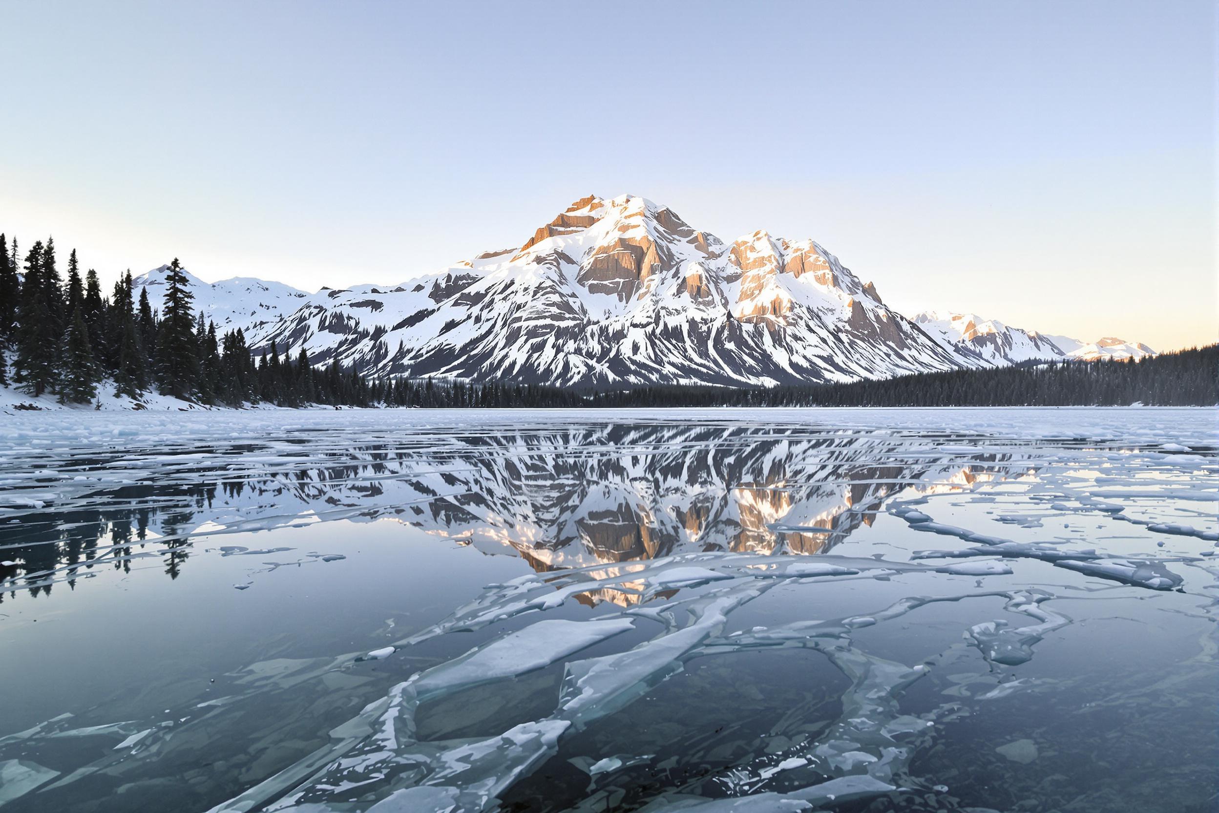 A pristine alpine lake mirrors a snow-capped mountain under soft morning light. Layers of ice frame the water's edges in intricate patterns, while evergreen trees dot the distant shore. The clear, pale blue sky fades into subtle gradients, amplifying the serene atmosphere of the untouched wilderness.