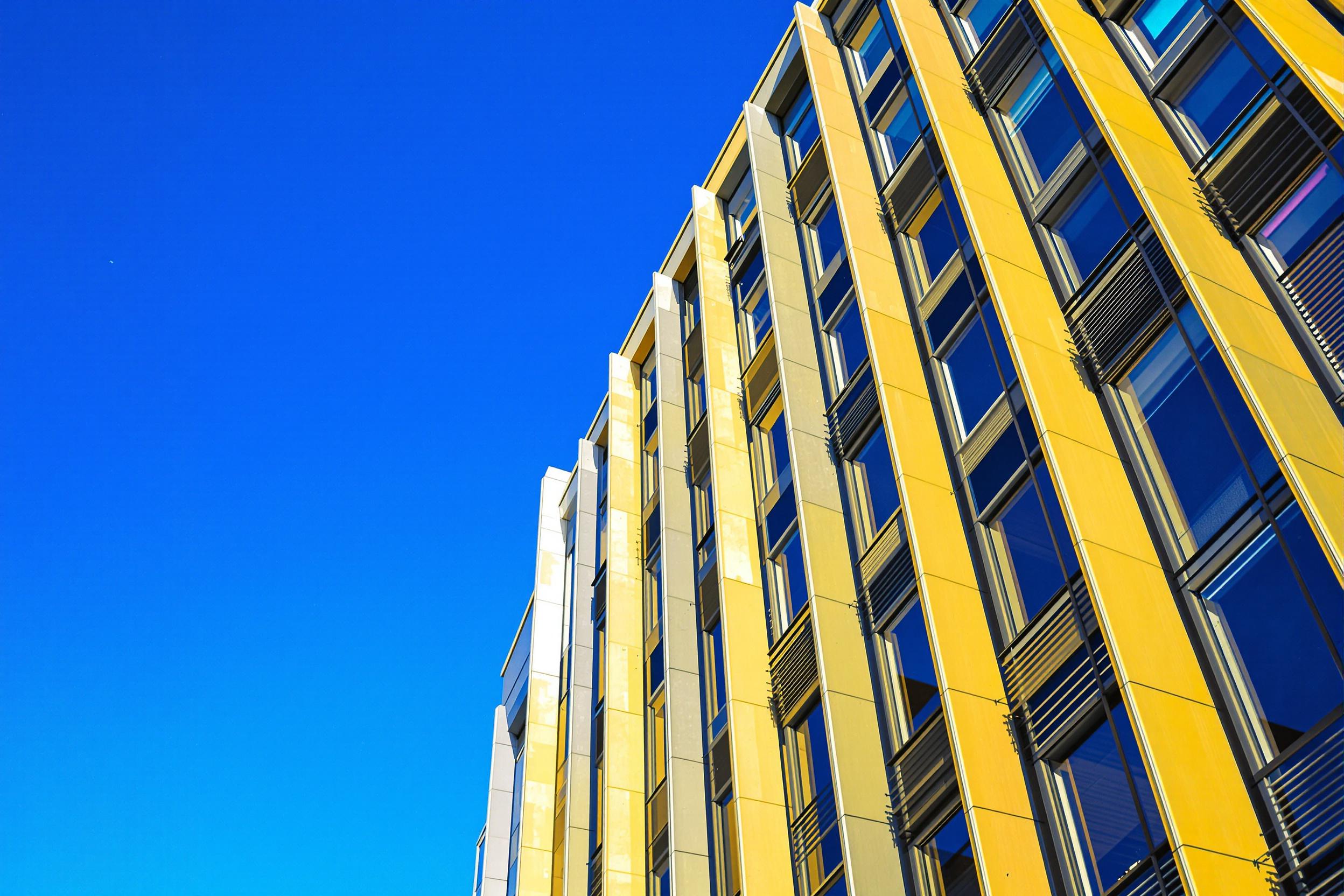 This striking image captures a modern urban building featuring bold geometric patterns and sleek lines. The composition highlights the interplay of light and shadow, with a vibrant blue sky serving as the backdrop. The warm sunlight enhances the building's textures, creating a dynamic visual experience. Ideal for architectural design and urban planning projects.