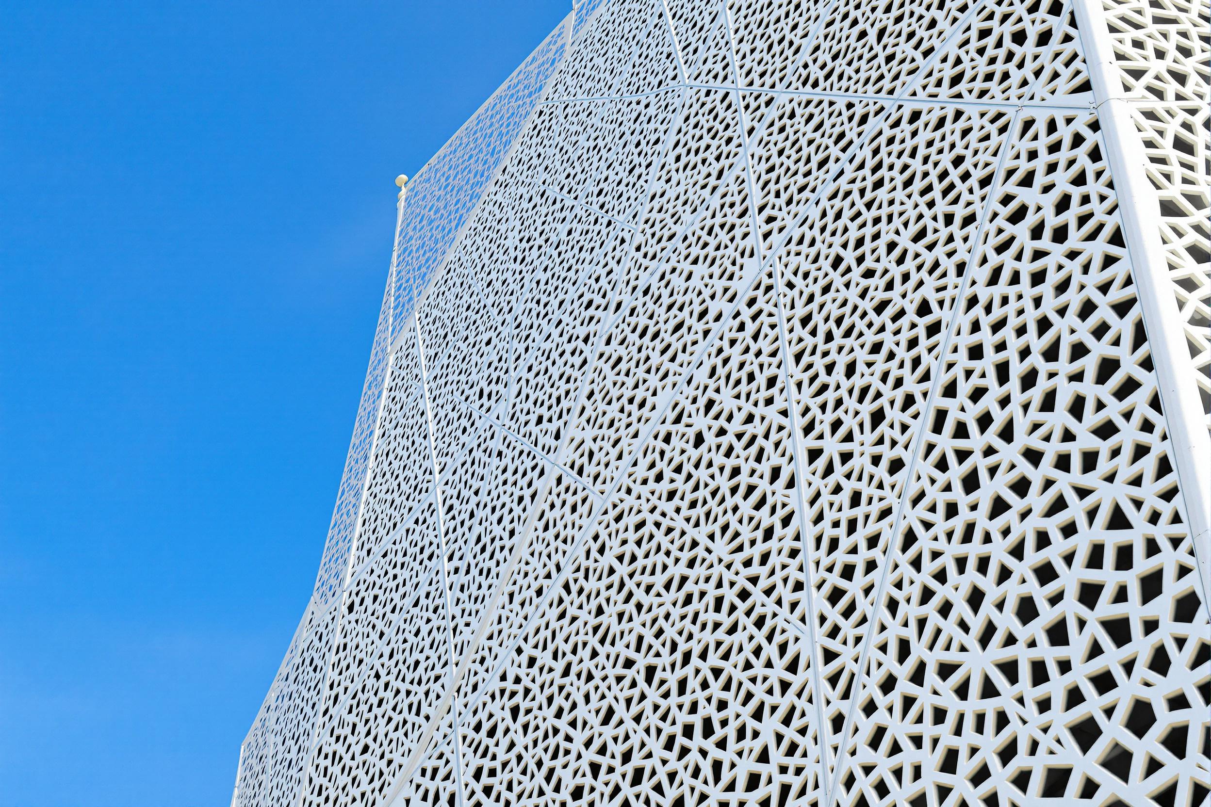 Close-up view of intricate geometric patterns adorning the exterior of a contemporary mosque. The striking white latticework contrasts beautifully against the azure sky, showcasing the seamless blend of traditional Islamic design elements with modern architectural techniques.