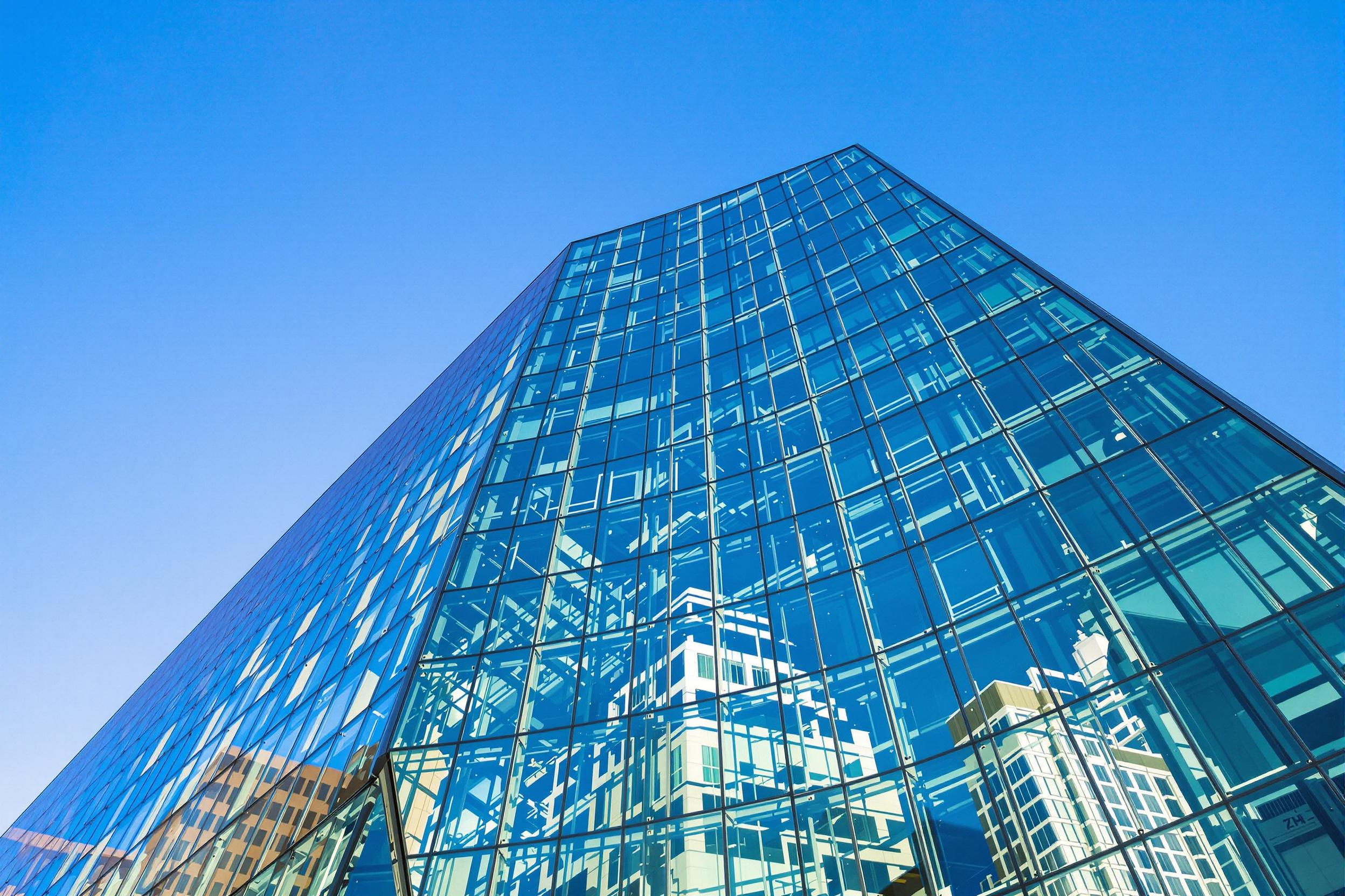 A striking image of a modern office building featuring a sleek glass facade that reflects the surrounding cityscape. The composition captures the building's angular lines and contemporary design, set against a clear blue sky. The warm sunlight enhances the glass's reflective quality, creating a vibrant atmosphere. Ideal for business and architecture themes.