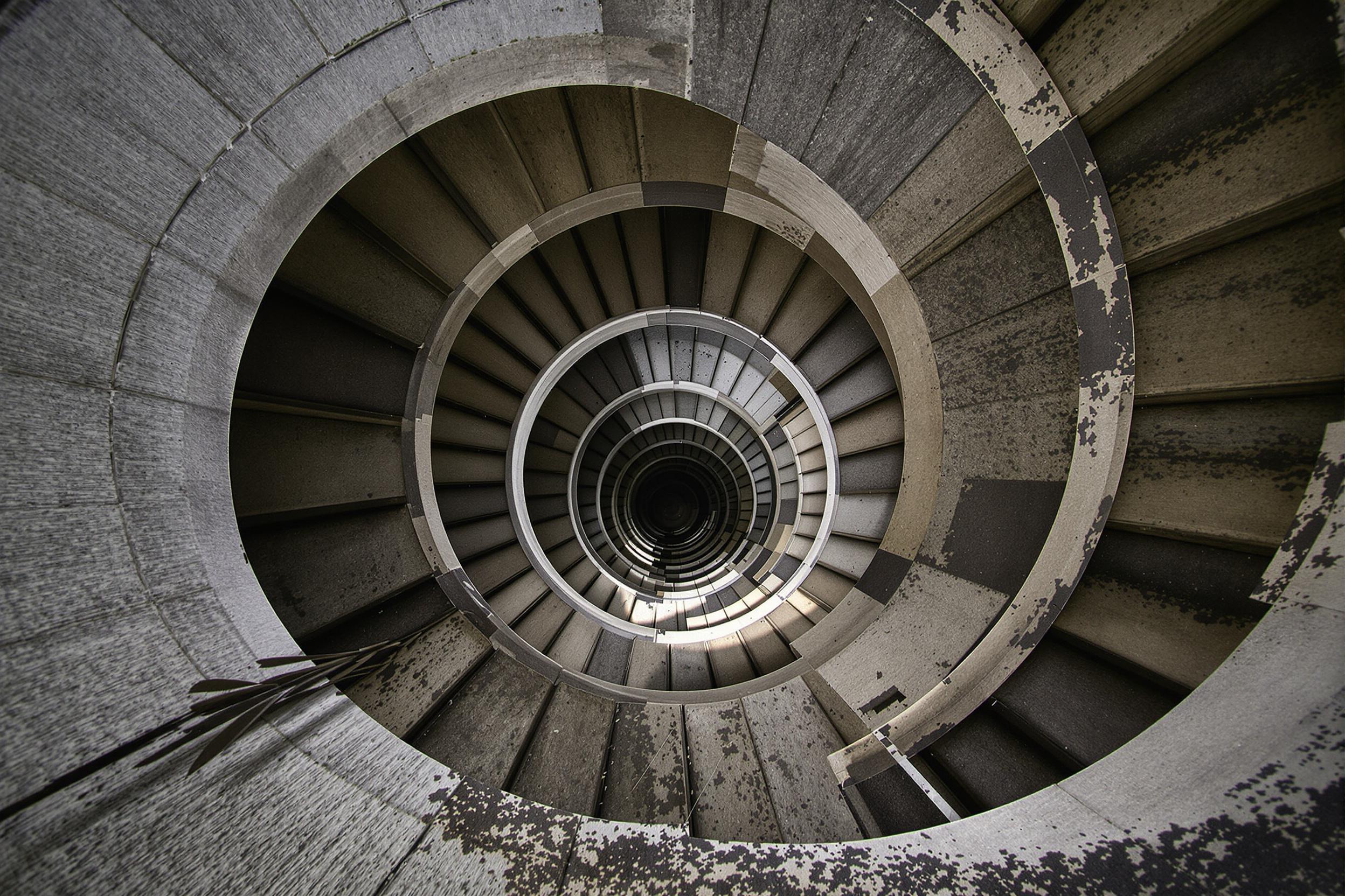 Striking image of a spiral concrete staircase in an abandoned industrial building. Raw concrete surfaces, dramatic shadows, and geometric patterns create a compelling composition that showcases the beauty of brutalist architecture and urban decay.