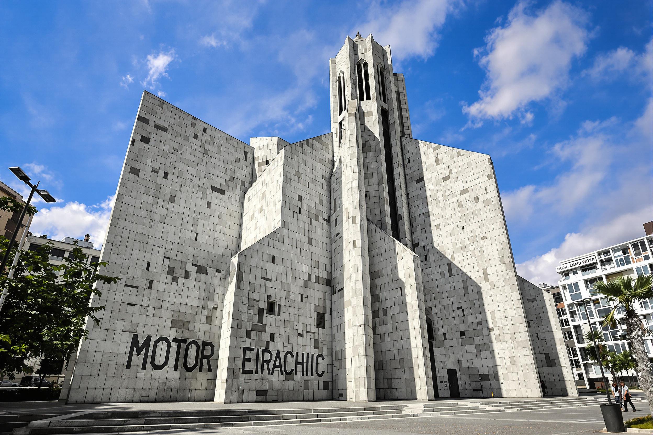 Discover a striking brutalist church rising from an urban landscape. This architectural masterpiece features raw concrete forms, dramatic angles, and a minimalist bell tower. Sunlight casts intricate shadows across the textured surfaces, creating a powerful interplay of light and shadow.