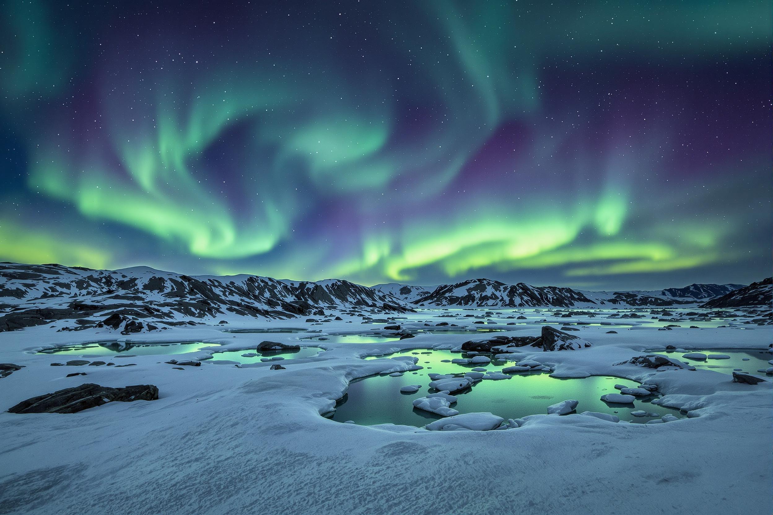 A frozen tundra stretches endlessly under a swirling canopy of the Aurora Borealis. The glacial terrain is illuminated by vivid green and purple lights, casting shimmering reflections on scattered ice pools. Jagged stone formations accentuate the dramatic expanse under the dark polar sky. Subtle texture on snow lends depth to the stark wilderness.