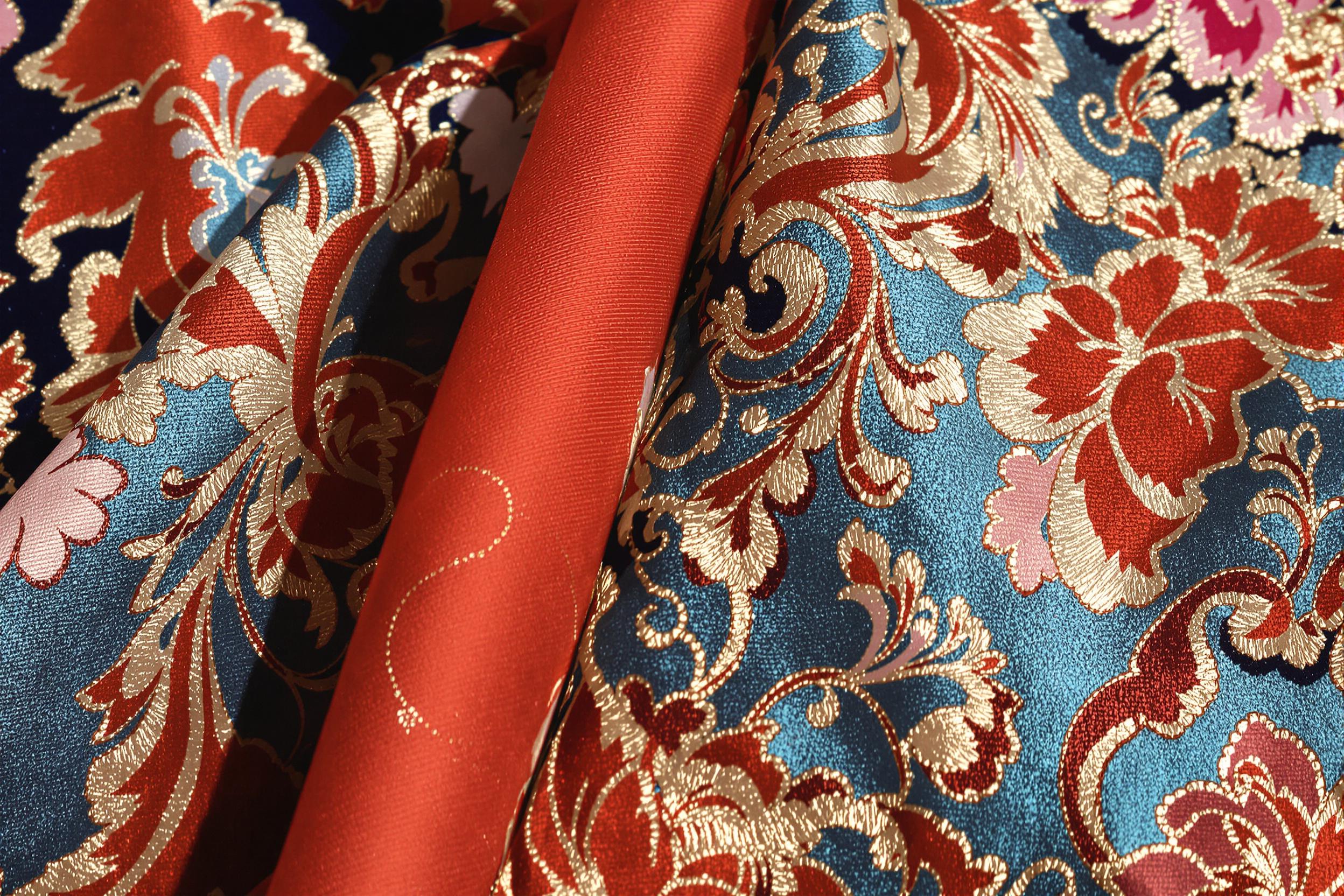 Extreme close-up detail of an intricately patterned kimono displays fine silk threads woven into floral designs with accents of gold embroidery. Hues ranging from deep reds and vibrant blues softly shimmer under diffused overhead light. The silky surface contrasts against subtle shadows from folds, adding depth to the image.