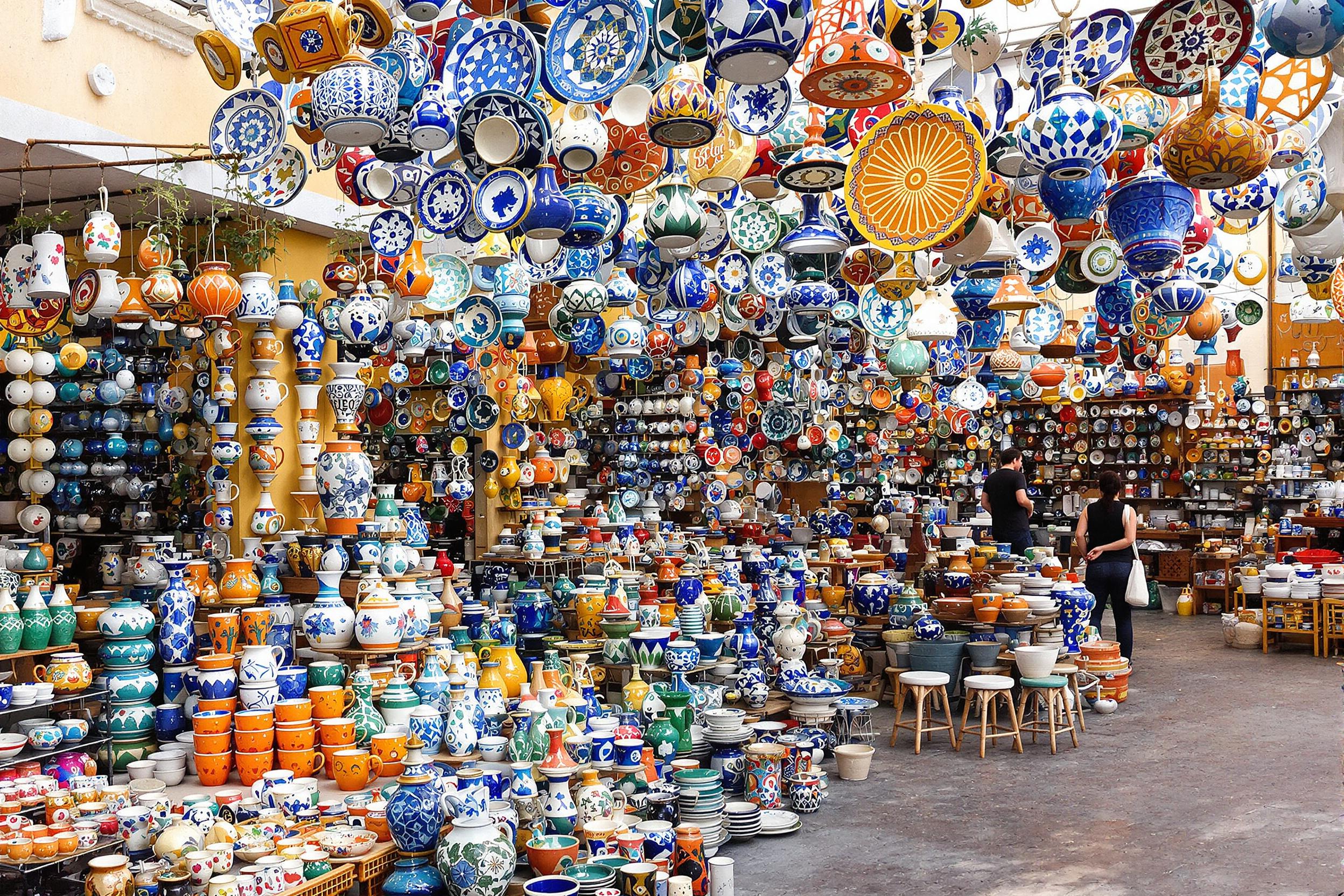 A vibrant open-air market bustles with artistic displays of handcrafted pottery. Colorful ceramics featuring intricate patterns catch the midday sunlight, creating a vivid mosaic of blue, orange, and green hues. Stalls brim with mugs, vases, and plates, while nearby artisans engage in their craft, inviting exploration of these unique creations.