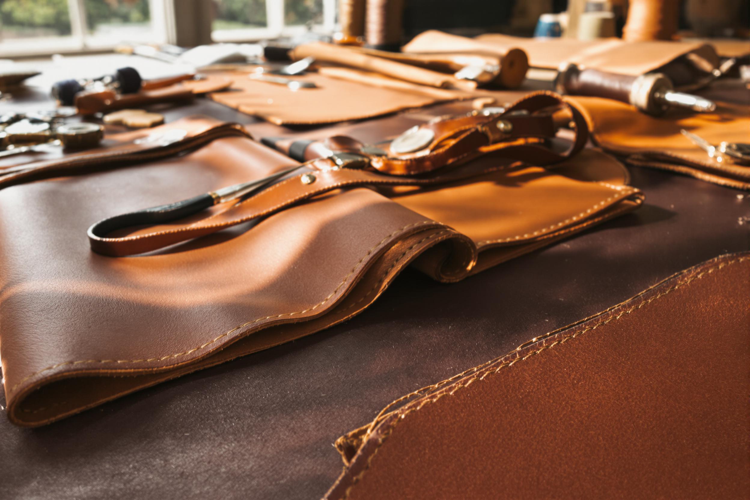 A close-up captures the meticulous craftsmanship of hand-stitched leather goods laid out on an artisan's workbench. Deep brown and caramel leathers reveal intricate stitching patterns, while tools like needles and thread complement the scene. Soft midday sunlight filters through nearby windows, enhancing rich textures and casting gentle shadows over this inviting workspace.