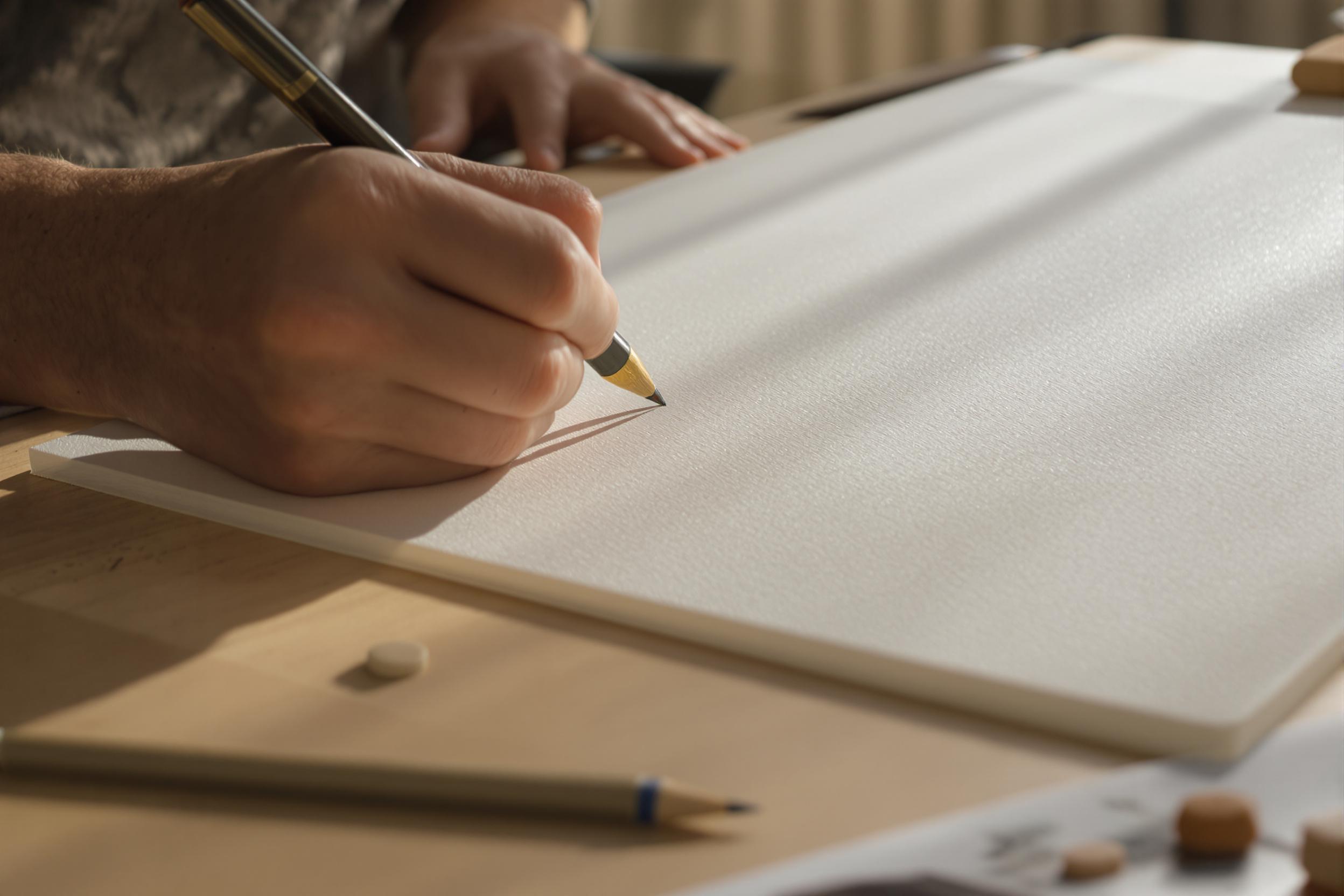 A close-up of an artist’s hand delicately sketching on a blank canvas. Soft, natural light streams through a nearby window, casting gentle shadows on the textured surface. The scene shows finely detailed pencils and erasers scattered on a maple-wood desk below the artwork, with everything bathed in muted beige and cream tones.