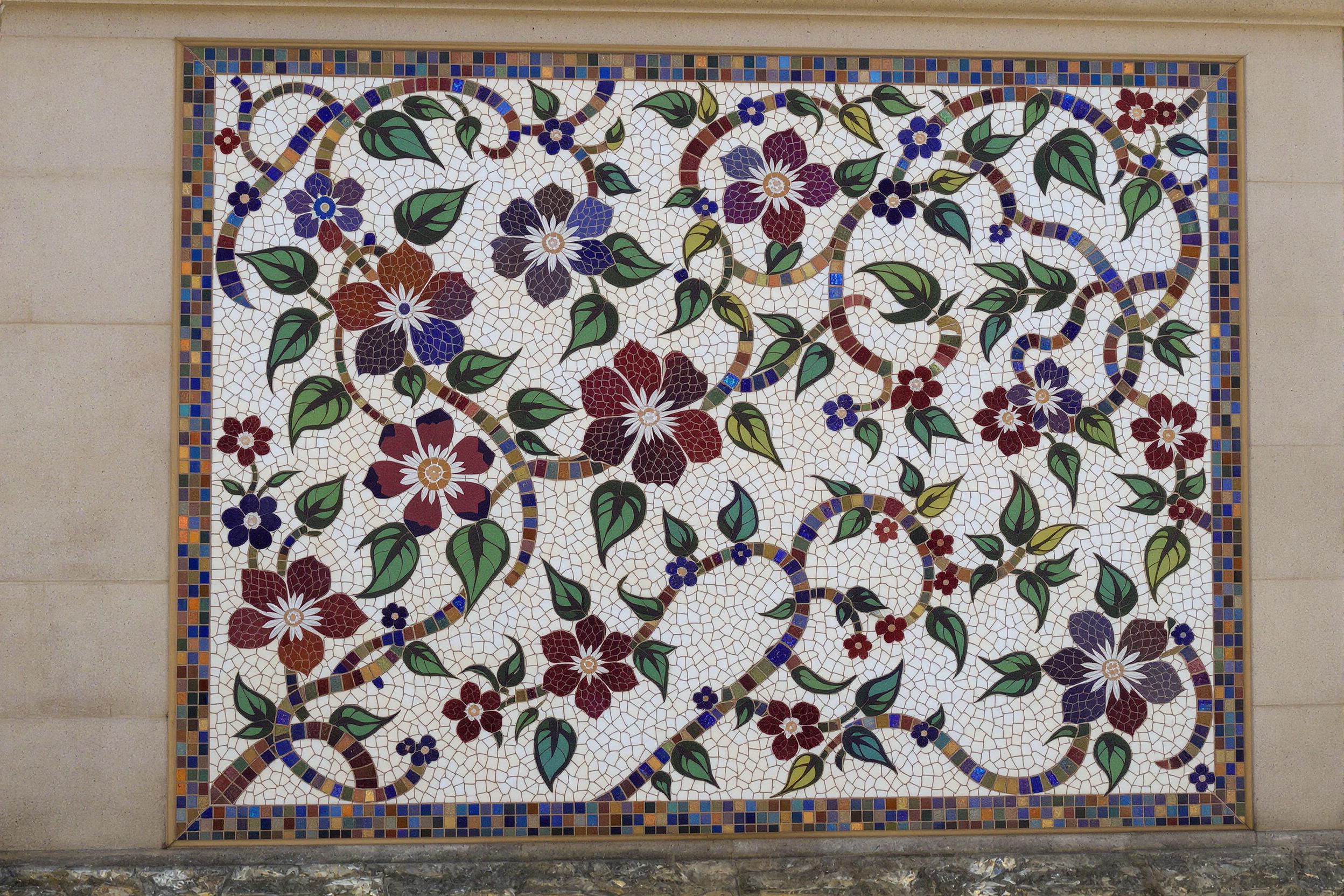 An intricate mosaic mural showcases a vivid depiction of blooming flowers intertwined with vines and leaves. Each piece of colored glass tile, ranging from deep blues and ruby reds to softer pastel tones, catches glints of sunlight. Set against a neutral stone wall, the shimmering artwork exudes vibrancy while evoking timeless craftsmanship.