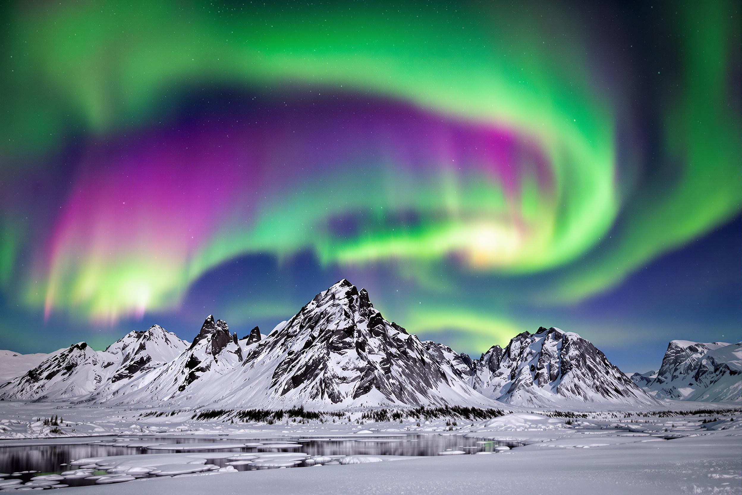 A breathtaking winter scene features snow-covered mountains under the dazzling aurora borealis. The colorful northern lights—green, pink, and violet—dance across the dark sky, creating undulating waves of light above rugged snow-capped peaks. Reflections shimmer subtly on icy terrain below amid the clear, serene Arctic atmosphere.