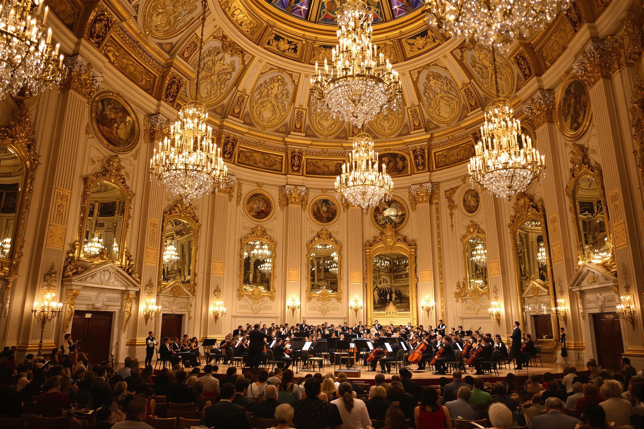 A grand baroque-style concert hall glows under elaborate crystal chandeliers. A small orchestra dressed in period-accurate attire performs for an attentive audience. Gilded ceilings and intricate stucco detailing contrast with polished mahogany flooring. The scene showcases soft, warm light bouncing off ornate wall mirrors.