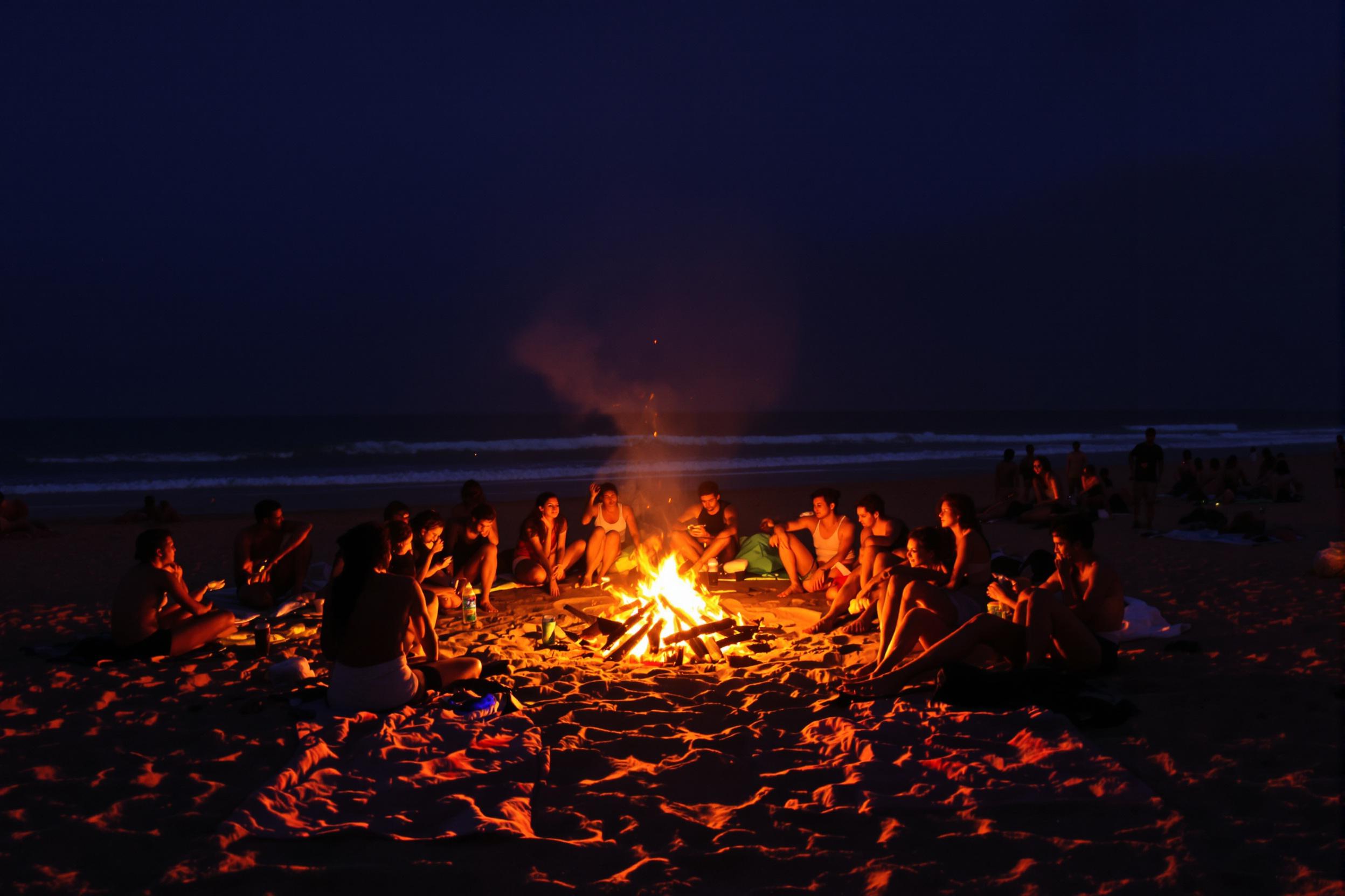 A wide-angle view captures a lively beach bonfire gathering as dusk settles in. Groups of friends sit close to the flickering flames, their faces illuminated by the warm glow. Wisps of smoke rise against a backdrop of deepening indigo sky, while soft waves lap gently at the shore. Colorful blankets are spread out, adding texture to the sandy surface, creating an inviting atmosphere for sunset conversations.