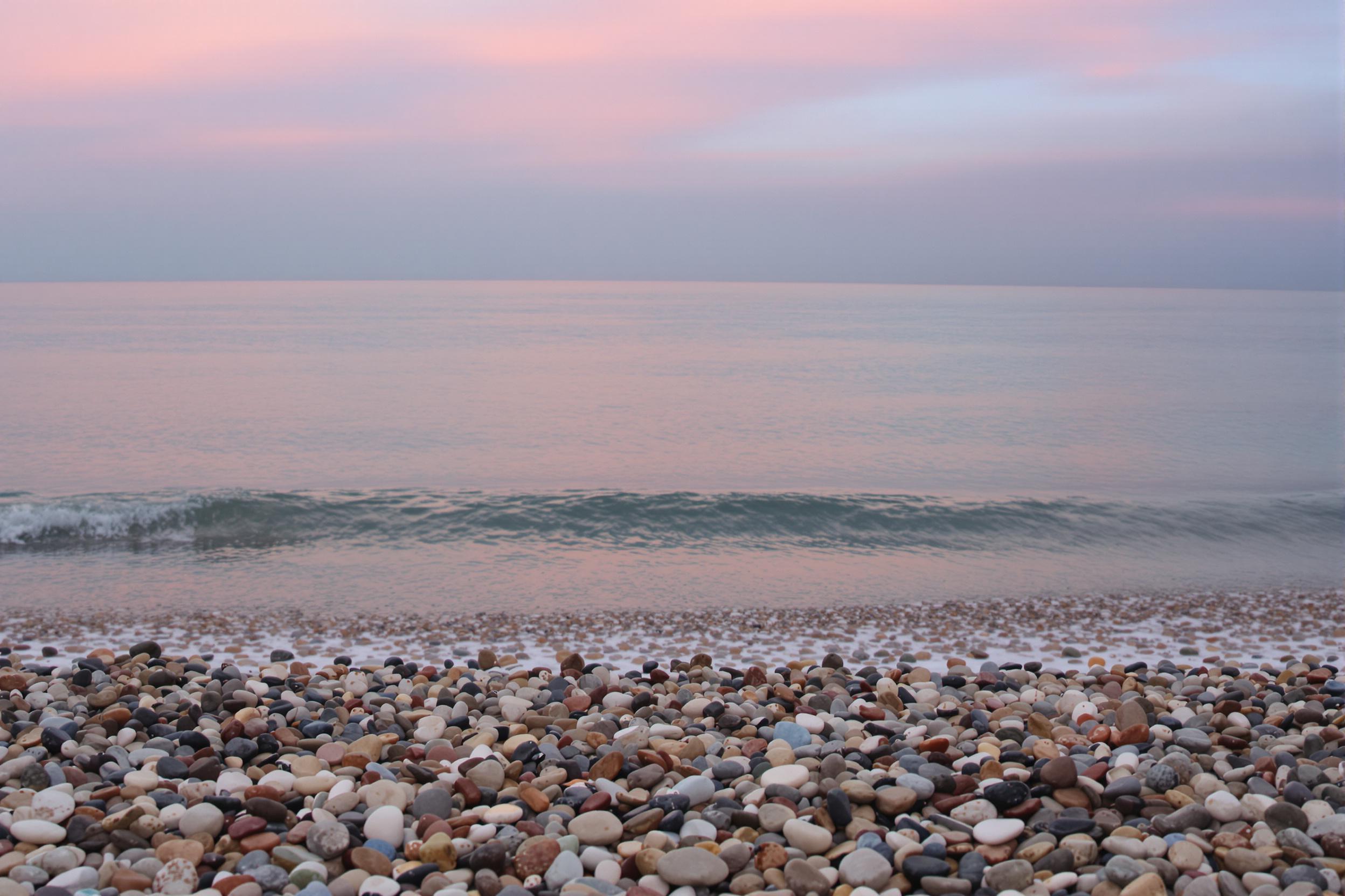 A tranquil beach scene unfolds at dawn, featuring a gentle shore lined with smooth, multi-colored pebbles. The calm sea reflects soft pastel hues from the awakening sky, blending pinks, purples, and blues. Light waves kiss the shoreline, creating delicate ripples. The serene atmosphere invites quiet reflection and appreciation for nature's beauty.