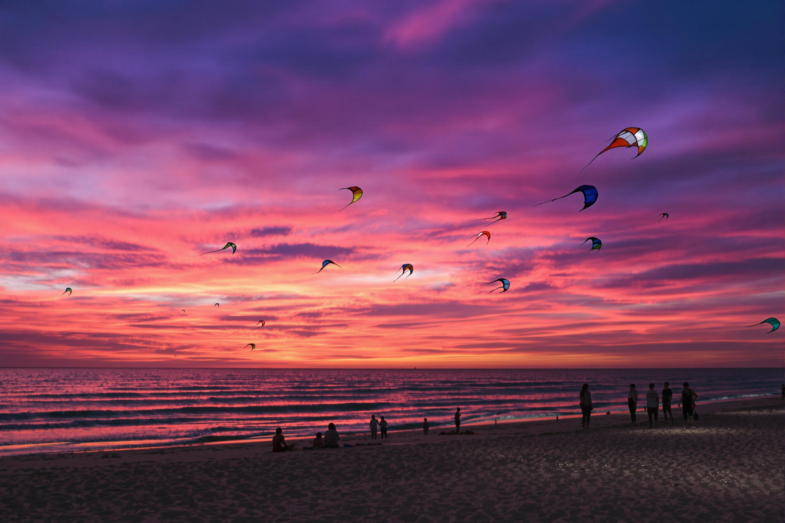 A breathtaking dusk beach scene captures colorful kites soaring against a vibrant sky. The horizon displays hues of purple, orange, and pink as the sun sets, reflecting off the calm ocean waters. Silhouetted figures stand scattered along the shore, their laughter mingling with the sound of gentle waves. Soft sands underfoot glisten in the twilight glow.