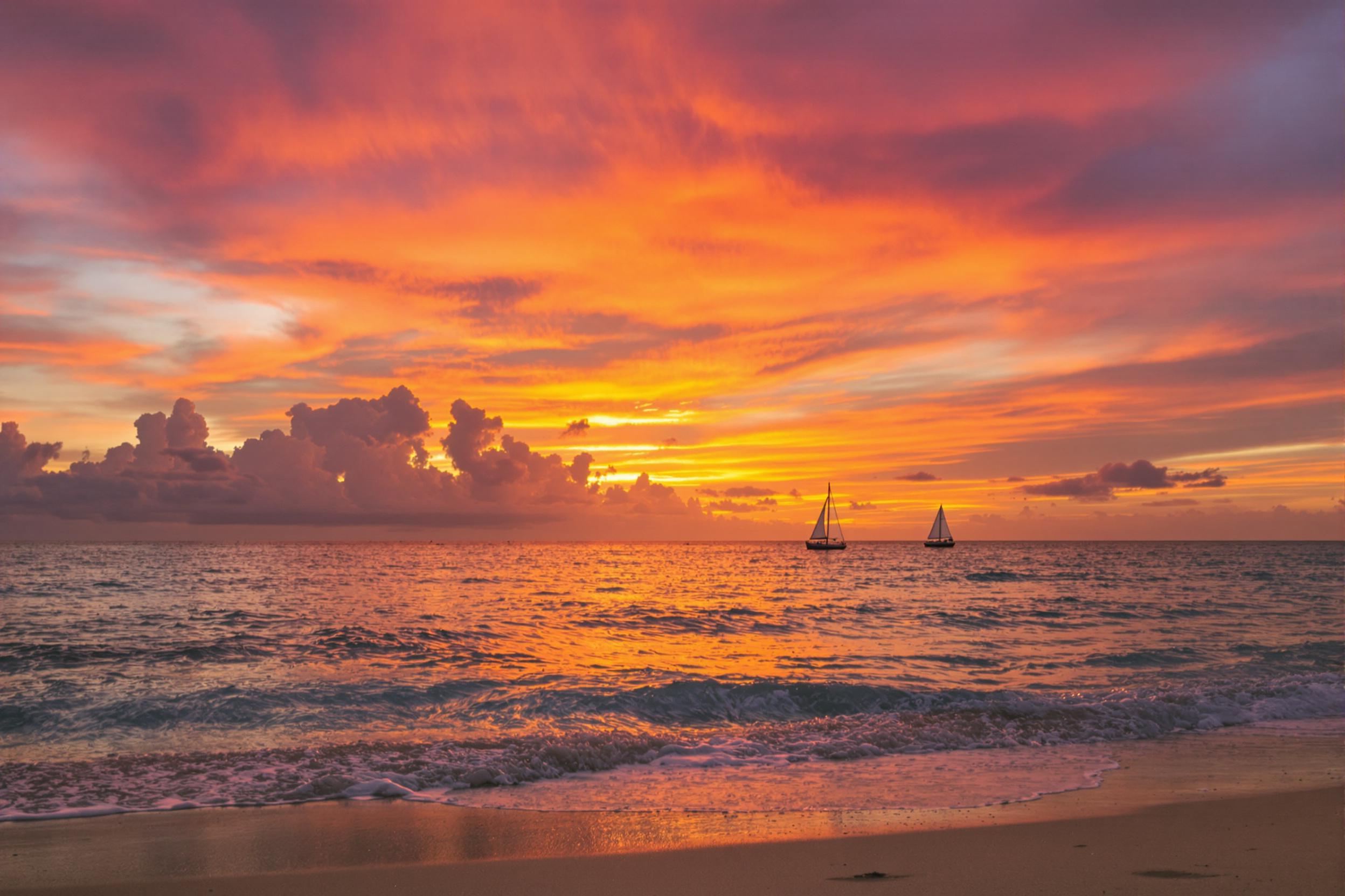 A serene beach scene unfolds at sunset, where gentle waves kiss the sandy shore. The sky is ablaze with vibrant orange, pink, and purple hues as the sun dips below the horizon. A solitary sailboat drifts peacefully on the glimmering water, its silhouette contrasting against the fiery sky. Silky sand glistens in the soft light, enhancing the tranquil ambiance of this idyllic panorama.