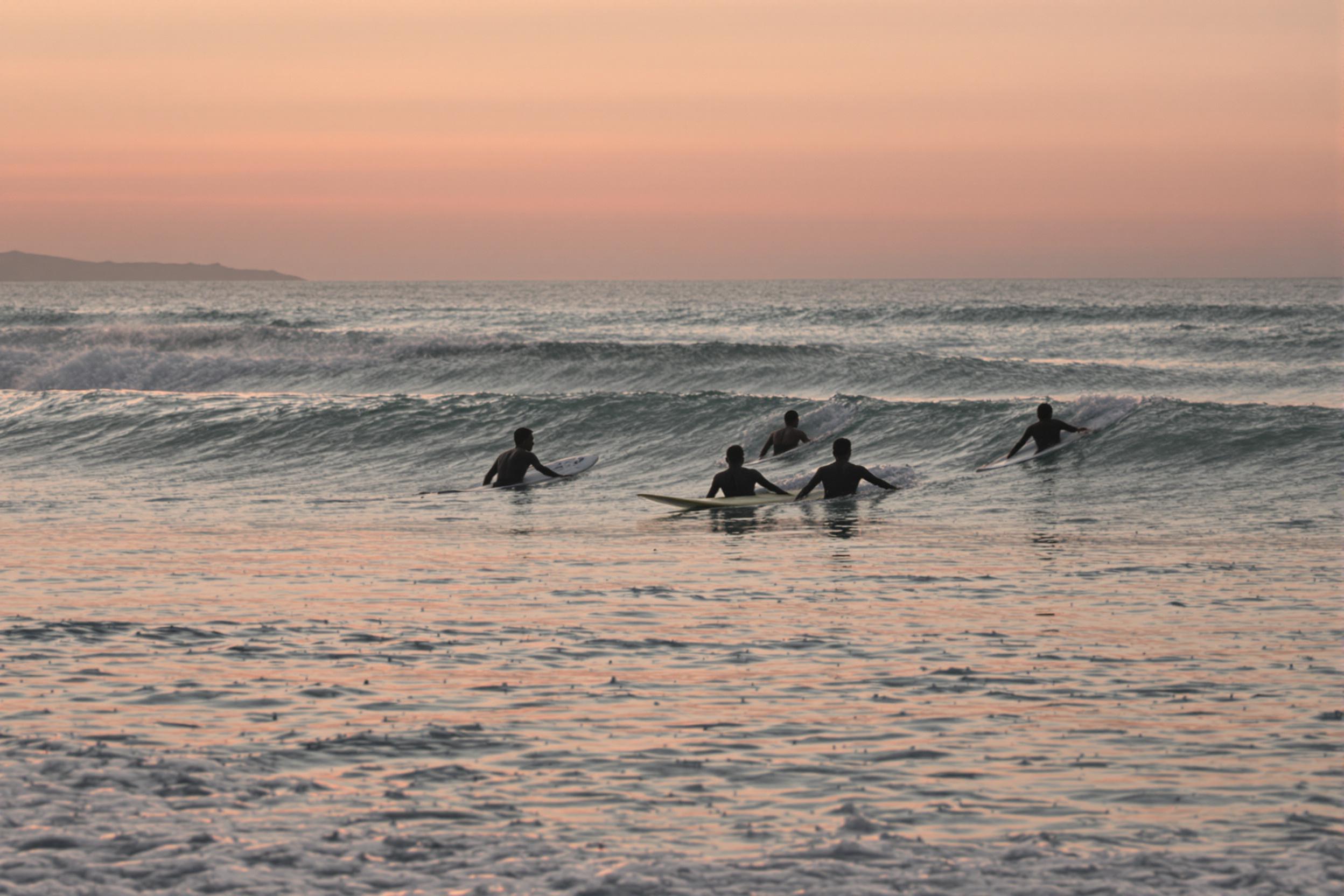 A tranquil beach scene unfolds as the sun sets, casting warm hues across the horizon. Silhouetted surfers gracefully navigate gentle waves, their outlines contrasting against the soft pink and orange sky. The subtly moving waters reflect the colors above, enhancing the serene ambiance of this picturesque coastal setting.