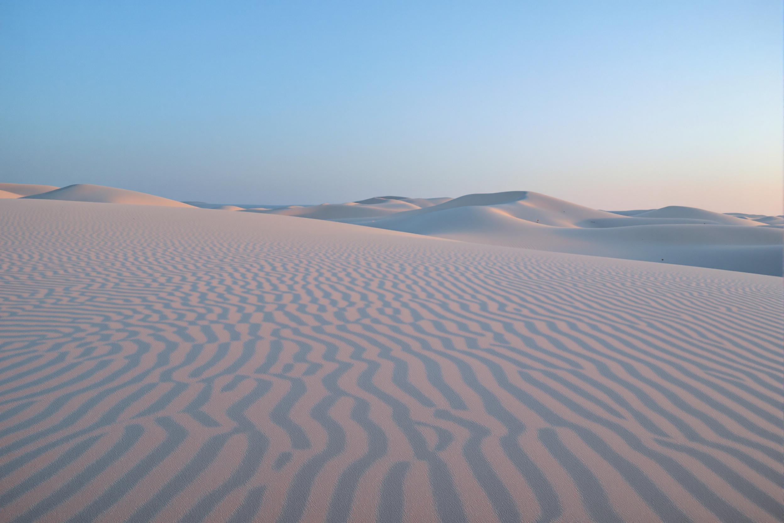 During the tranquil blue hour, endless rolling sand dunes stretch softly across the frame. Delicate curves catch traces of faint twilight, shifting between muted golds and deep cobalt blues. Fine surface ripples, cast in high detail, create intricate textures blending seamlessly into the soft, undulating horizon. The scene evokes dreamy solitude.