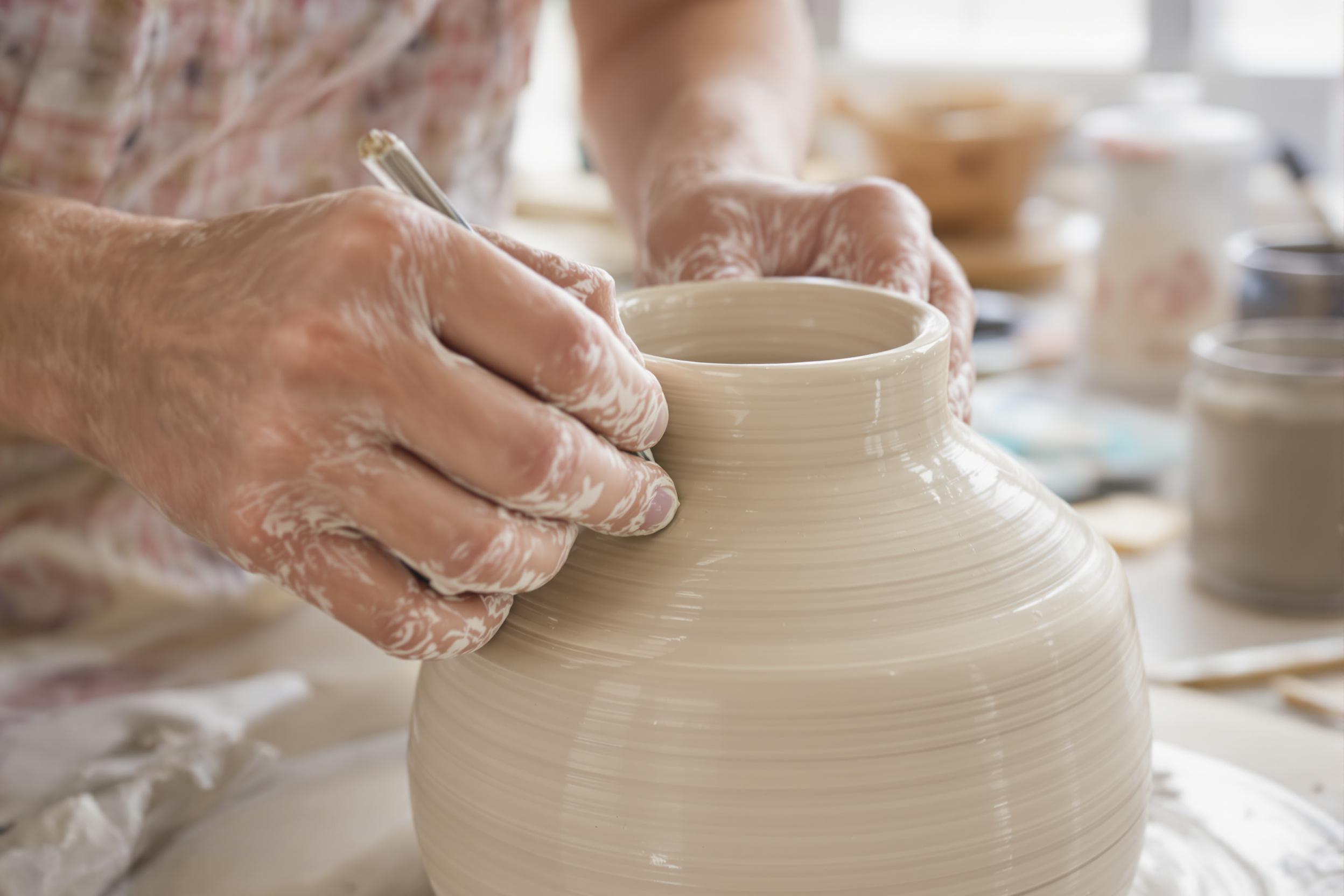 A close-up image captures the moment a potter’s steady hands apply glaze to a handcrafted ceramic vase. The soft daylight filters from a nearby window, enhancing the glossy texture of the glaze as it spreads evenly across the clay surface. Scattered tools and containers subtly fill the blurred background, completing this intimate artisan scene.