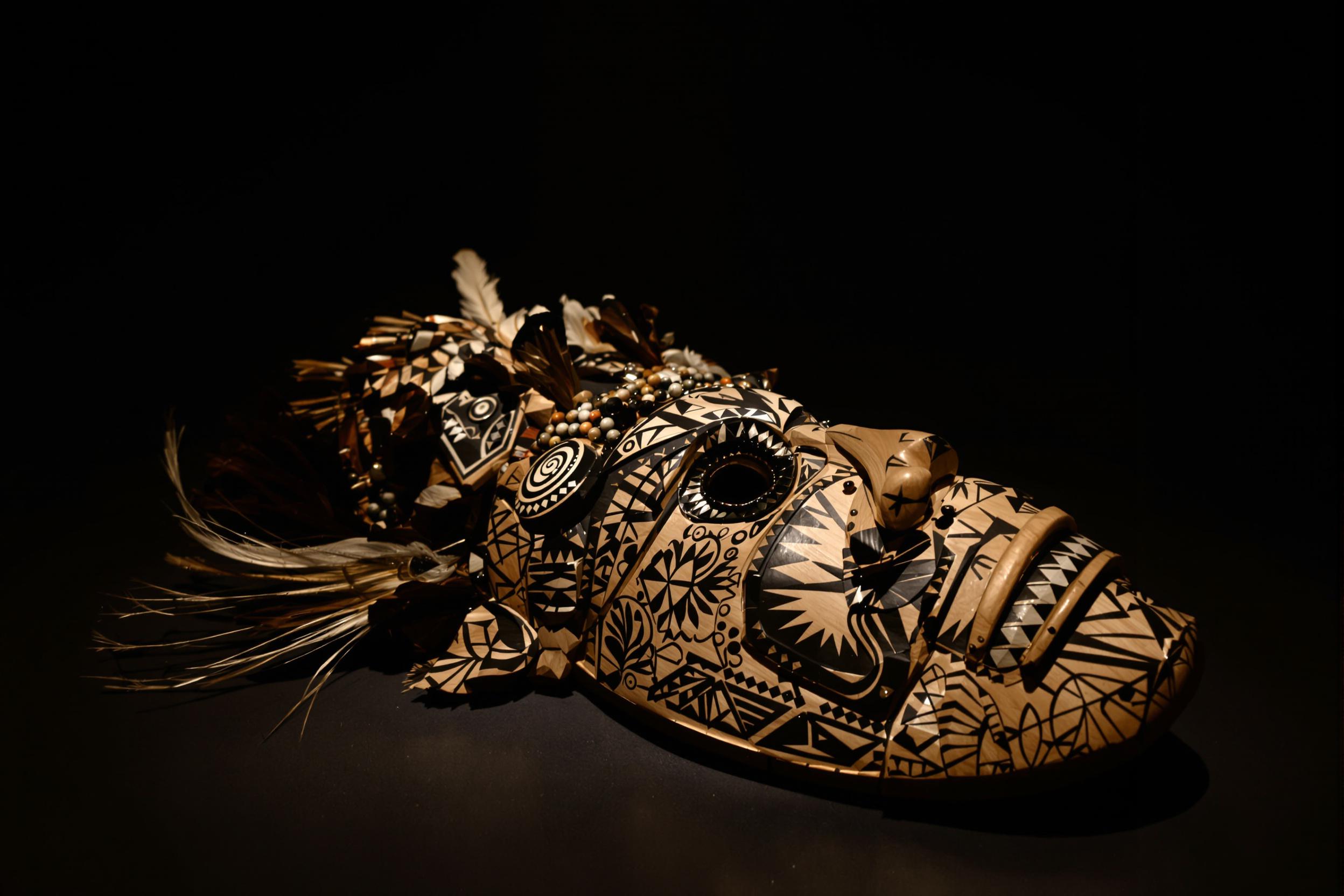 An intricately designed ceremonial mask rests bathed in soft, directional light within a quiet museum setting. The wooden mask features bold etched patterns adorned with beads, feathers, and fine metallic inlays. Surrounding shadows define its three-dimensional details, while a dark earthy backdrop focuses attention on its cultural artistry.