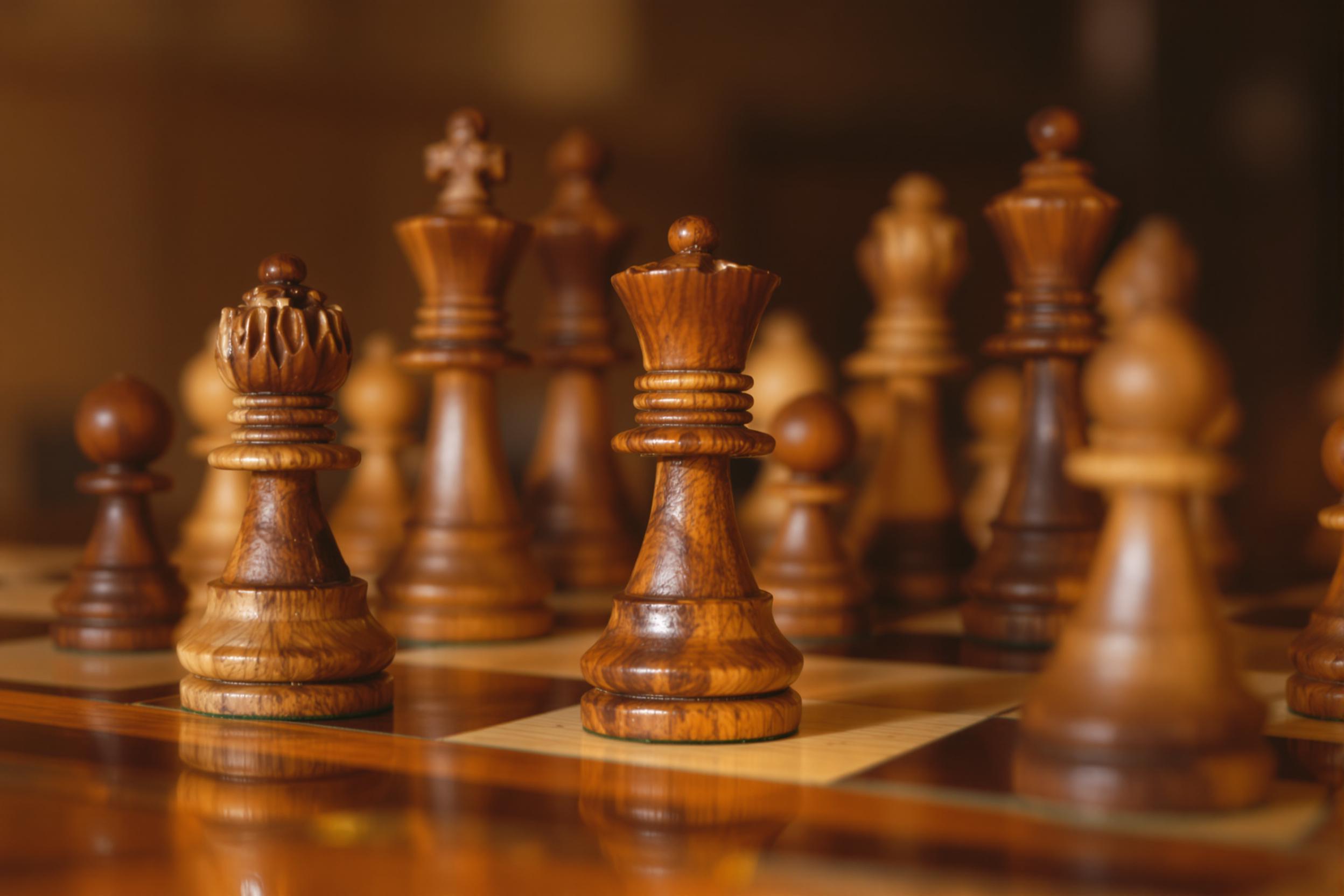 A close-up displays polished wooden chess pieces intricately hand-carved, positioned mid-game on an antique chessboard. Warm, directional light captures the rich wood grains and subtle wear on the aged surface. The blurred background fades softly, keeping focus on the meticulous detailing and tactical arrangement of the central pieces.