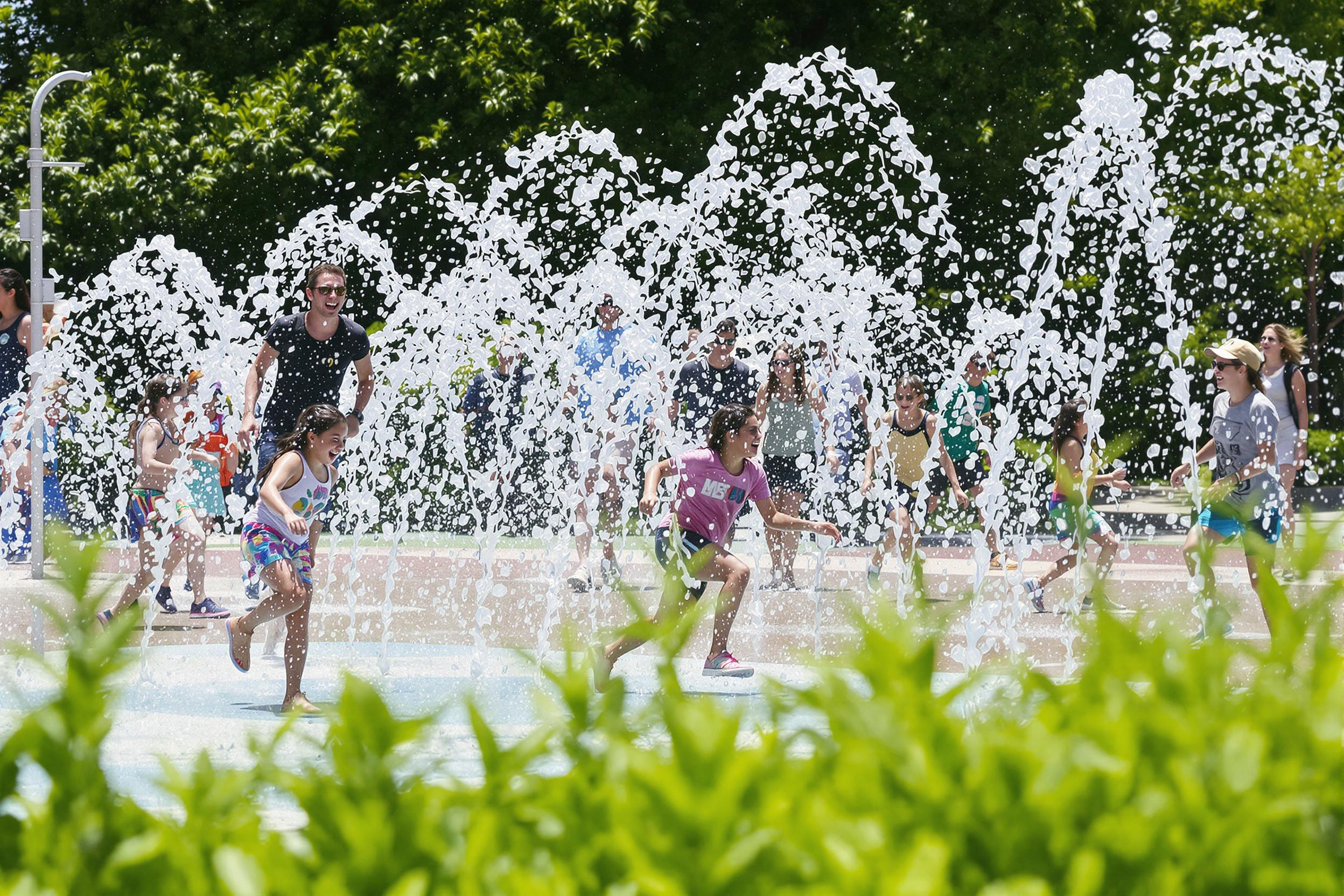 A lively splash pad bursts with joy as children dash through arcs of sparkling water under the midday sun. Laughter fills the air while droplets shimmer in the bright light. Surrounding lush greenery enhances the scene, creating a cheerful, playful atmosphere that celebrates summer fun.