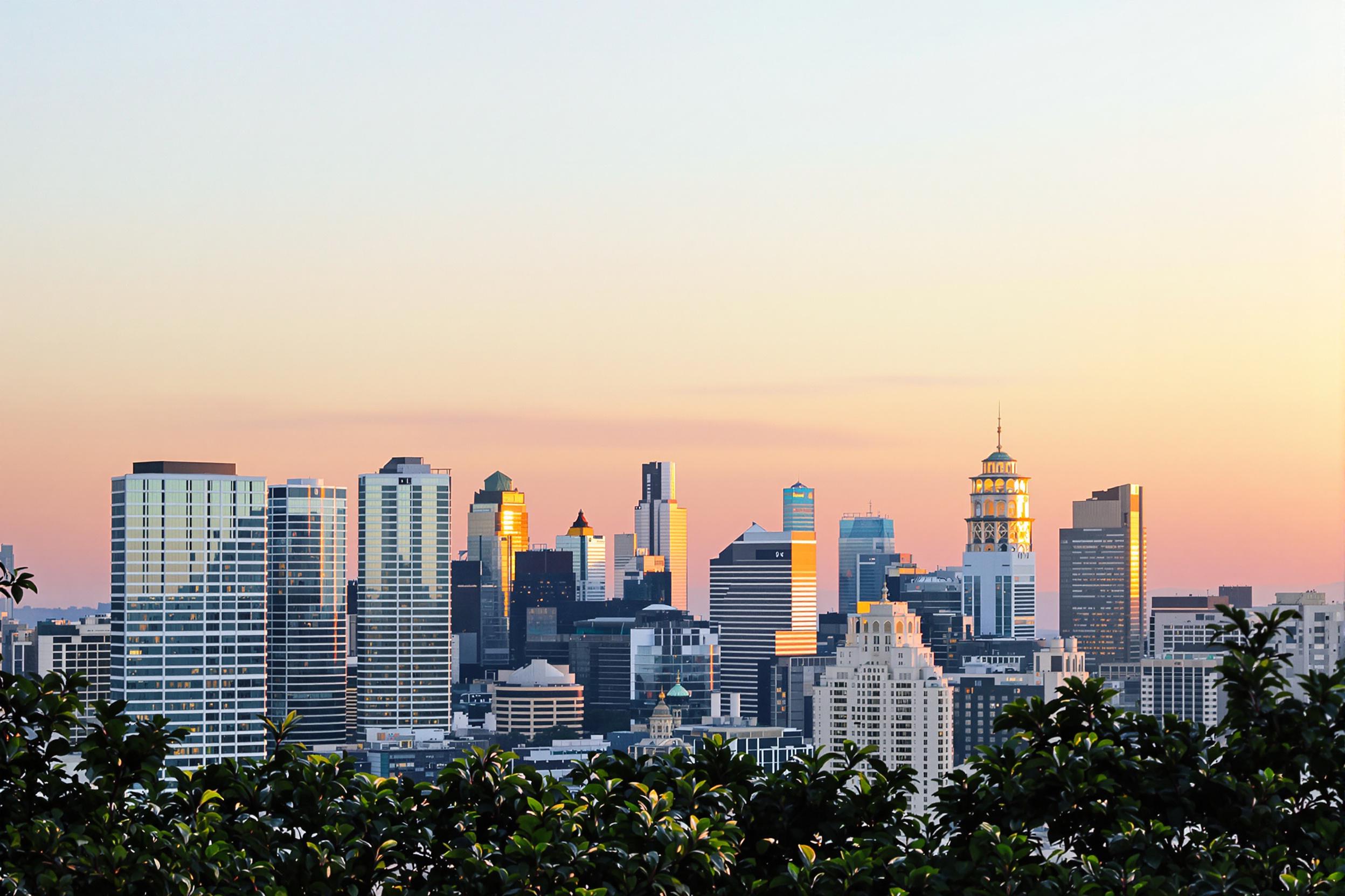 An expansive view of a vibrant city skyline at sunrise. Skyscrapers rise majestically against a backdrop of soft pastels as the sun begins to crest the horizon. Warm oranges and pinks illuminate the windows, creating a shimmering effect across the glass facades. Below, silhouettes of trees frame the scene, enhancing the urban landscape's charm.