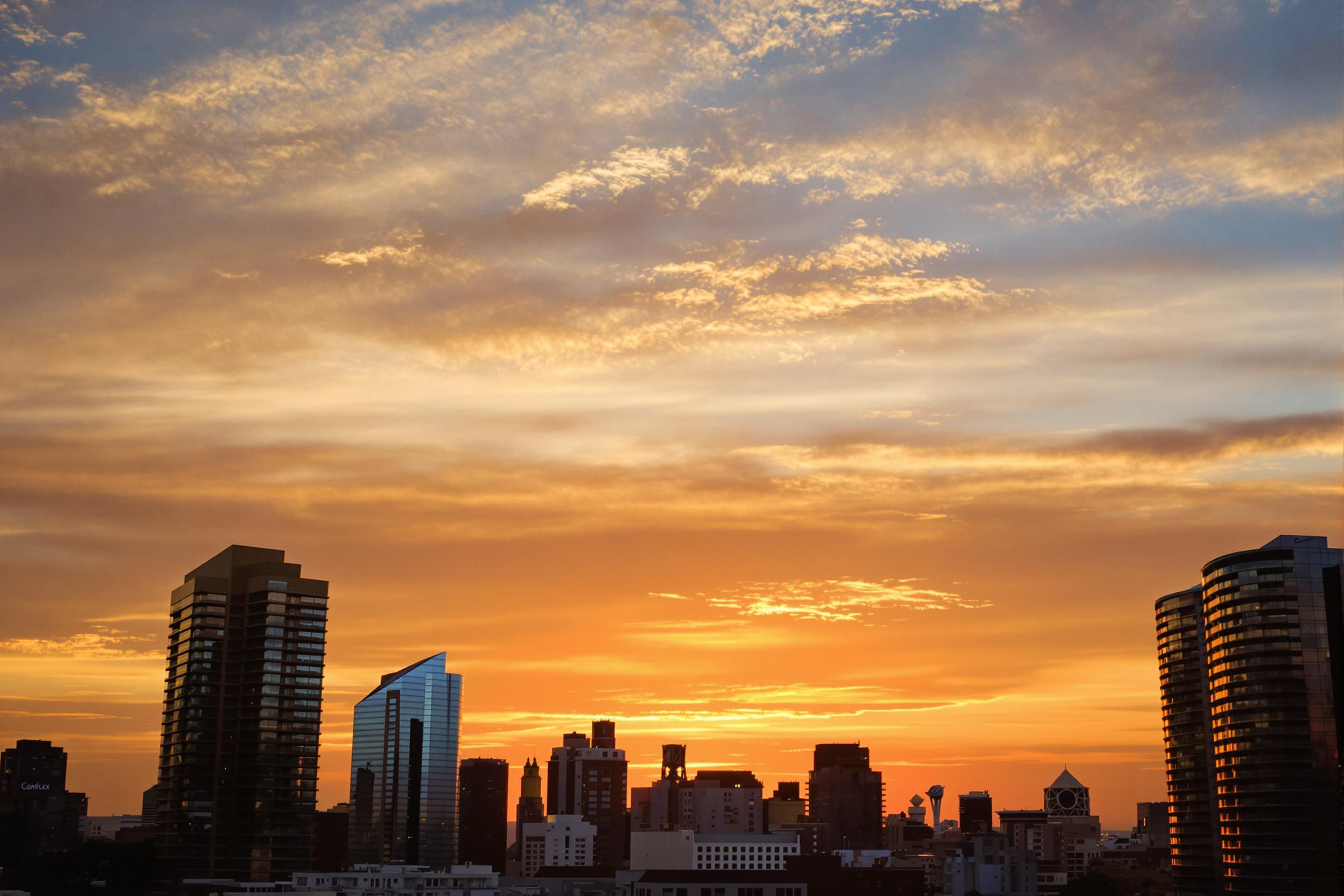 An enchanting sunset blankets a bustling city skyline. Golden, orange, and pink hues paint the sky as the sun dips below the horizon. Tall skyscrapers create striking silhouettes against the vibrant backdrop. Wispy clouds catch the light, adding texture. The scene conveys a sense of calm amidst urban life, marking the transition from day to night.