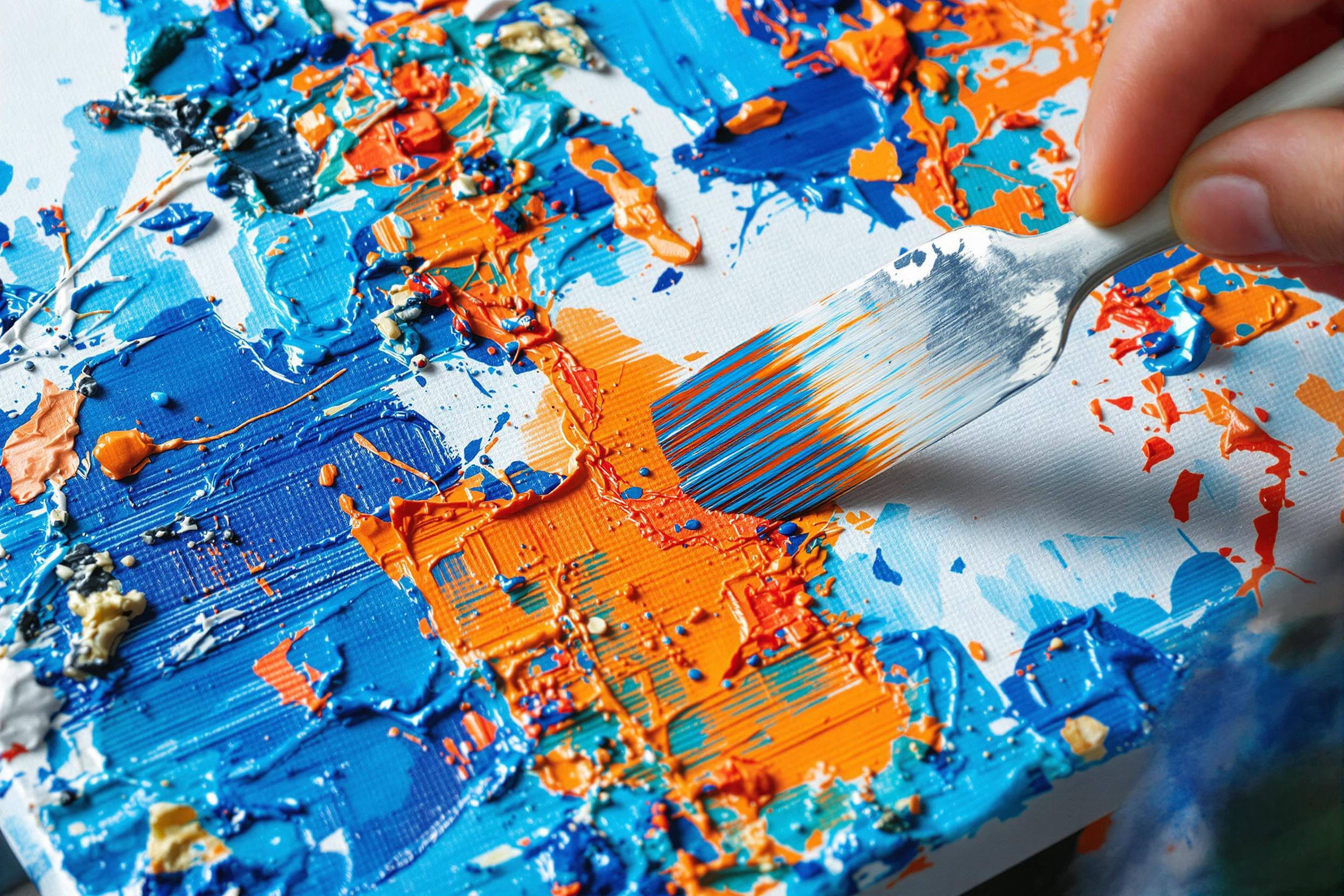 A textured view of an artist applying bright oil paints to a white canvas using a palette knife. The macro-level detail accentuates stacked colors—deep blues, fiery oranges, stark whites. Natural light enhances vivid hues and a dynamic, messy workspace surrounds suggesting spontaneity of abstract creation.