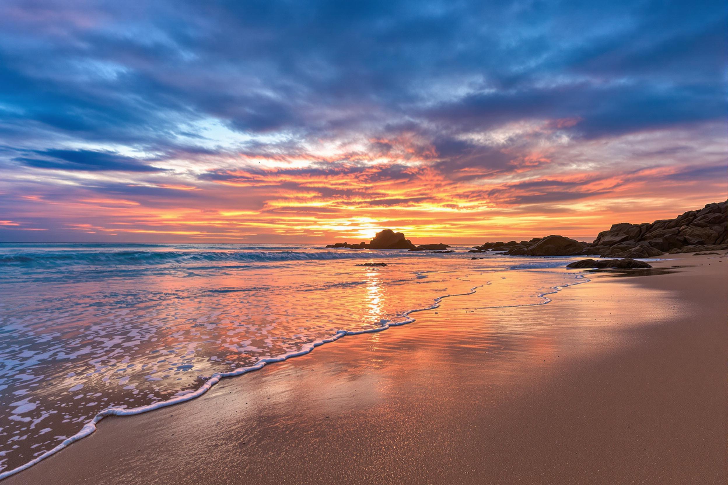 A serene coastal beach scene captures the gentle retreat of waves, leaving behind delicate tide lines gleaming in the gold-pink hues of a setting sun. Smooth sand reflects the colorful gradient sky, seamlessly blending deep blues and fiery oranges. Sparse rocky outcroppings add subtle contrast while creating focal points across the frame.