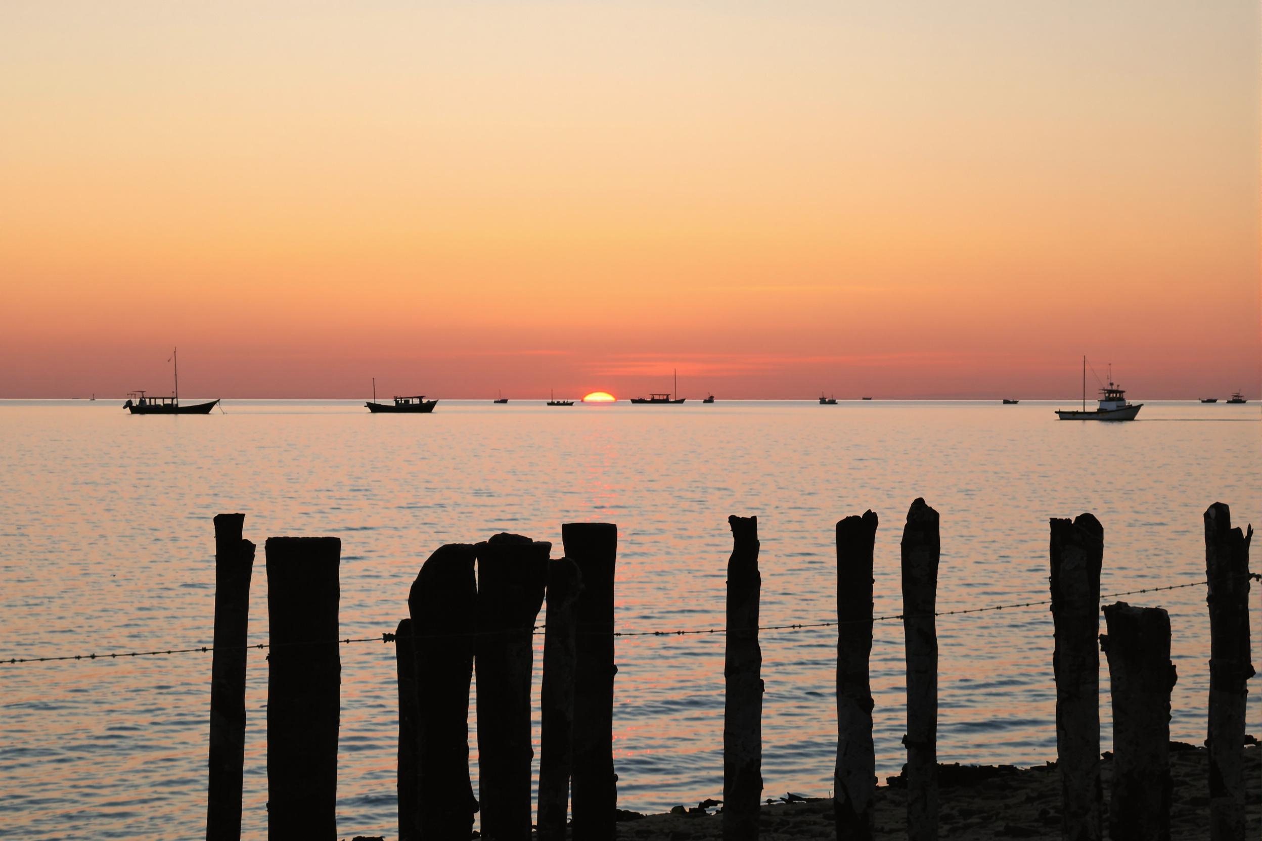 A tranquil coastal sunrise greets the morning as warm hues of orange and pink blend across the calm waters. Small fishing boats rest near the shore, their contours dark against the glowing horizon. Soft ripples on the water reflect the emerging sky above. The foreground reveals weathered wooden posts, framed naturally amidst this serene dawn moment.