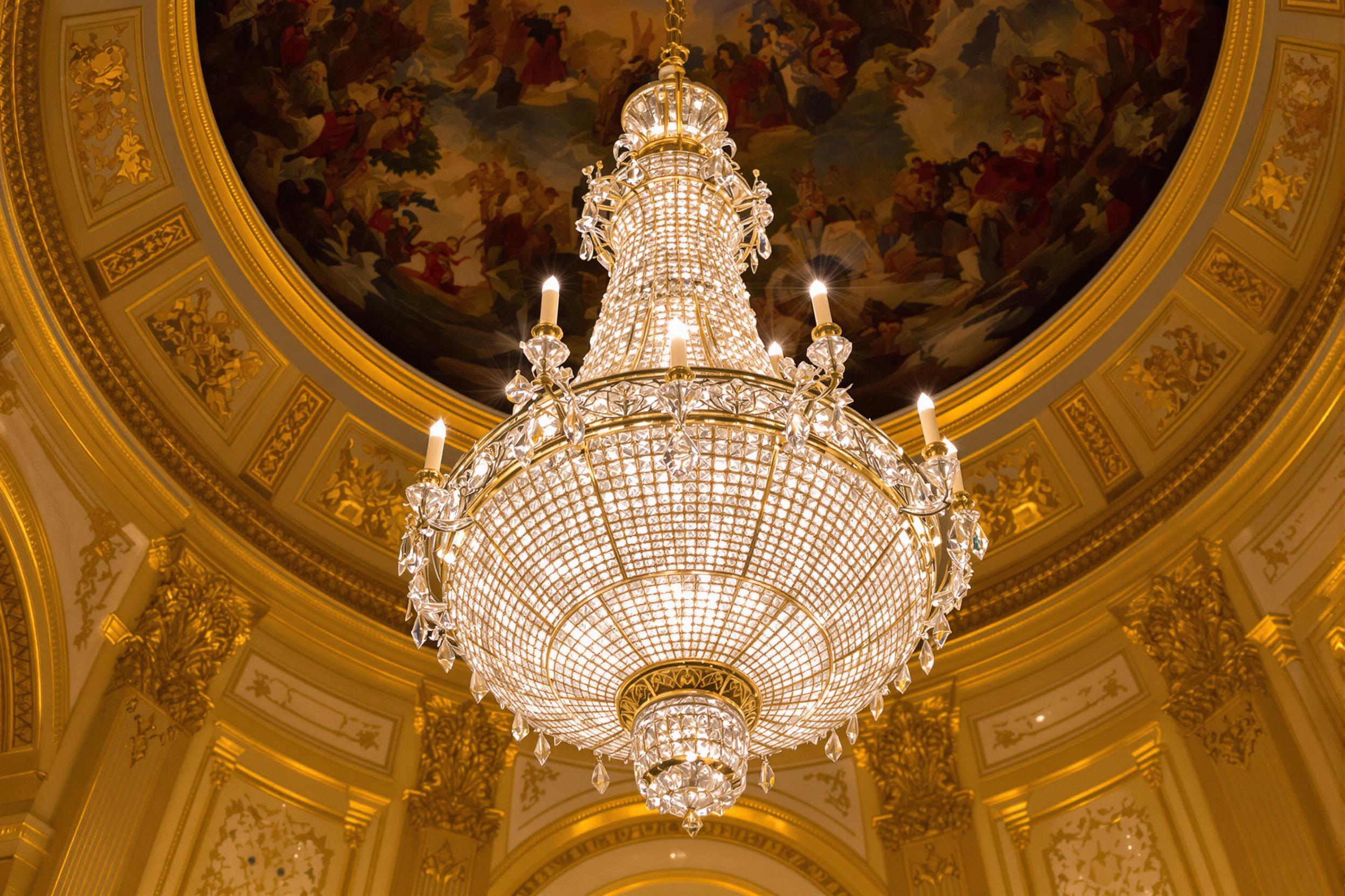 An aerial perspective captures a deeply ornate crystal chandelier suspended high in an opulent hall. The intricate glasswork glimmers as spotlights enhance its polished prisms, scattering faint radiant patterns onto surrounding gold-accented walls. Above, a frescoed dome ceiling is partially visible, framing this regal centerpiece.