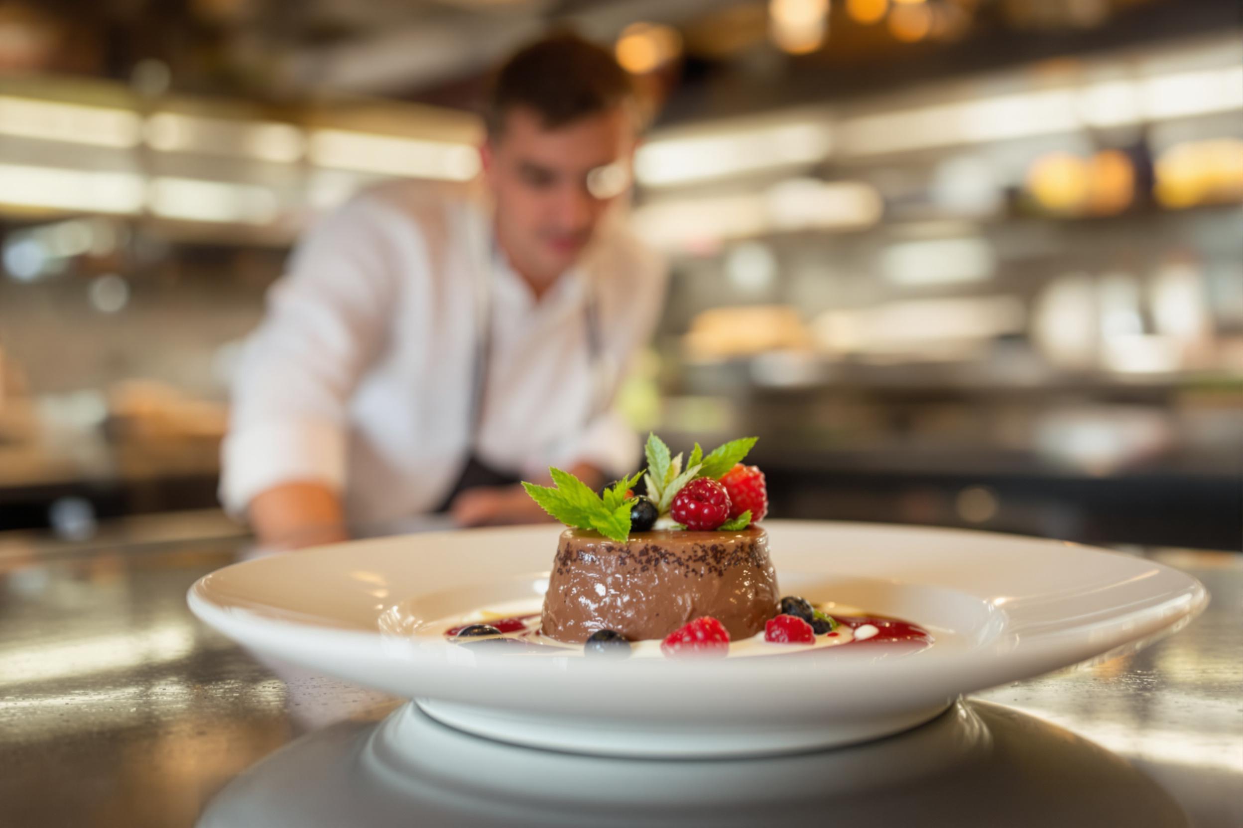 In a sophisticated restaurant kitchen, a skilled chef meticulously plates a stunning dessert. The dish showcases a rich chocolate mousse, artfully garnished with fresh berries and delicate mint leaves. Soft, warm lighting highlights the intricate details of the dessert, creating a luxurious presentation. The blurred background features polished countertops and culinary tools, emphasizing the artistry of fine dining.