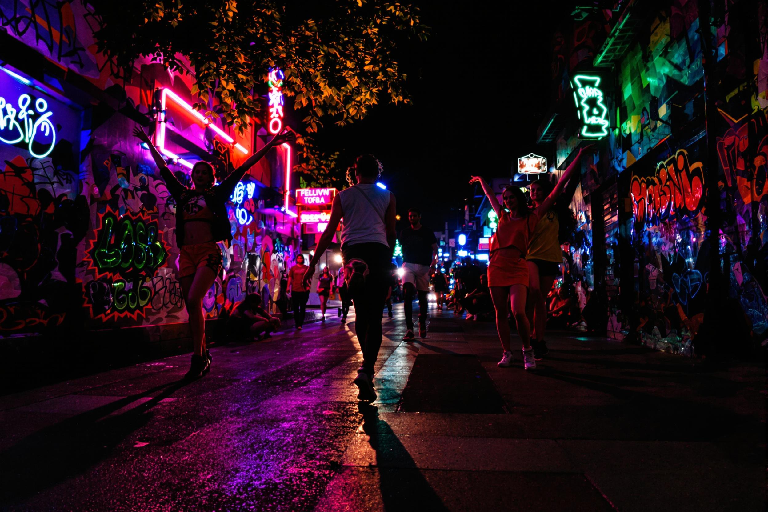 A captivating night scene captures a dynamic dance performance on an urban street. Neon lights illuminate the backdrop adorned with colorful graffiti, casting vibrant reflections onto the pavement. Dancers move gracefully, caught mid-motion, their colorful outfits contrasting against the dark ambiance. Shadows stretch and pulse with their movements, enhancing the electric energy of the atmosphere.