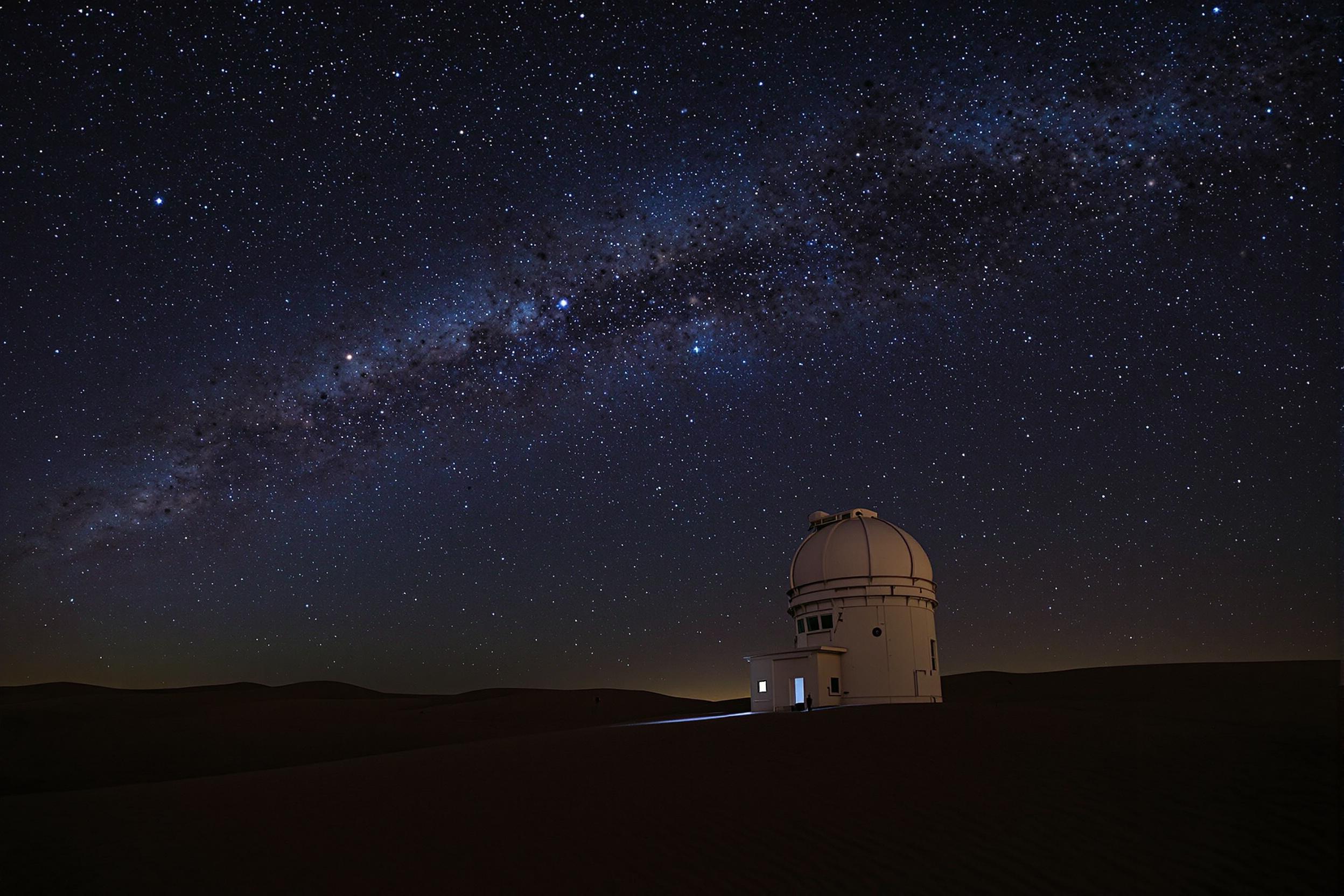 A remote observatory stands solitary on a sandy desert plain under a sweeping star-filled night sky. The faint, ethereal glow of the Milky Way arches high above. Soft blue light emanates from the dome, hinting at ongoing study. Shadows from rippling dunes provide subtle foreground textures, enhancing the sense of isolation and wonder.