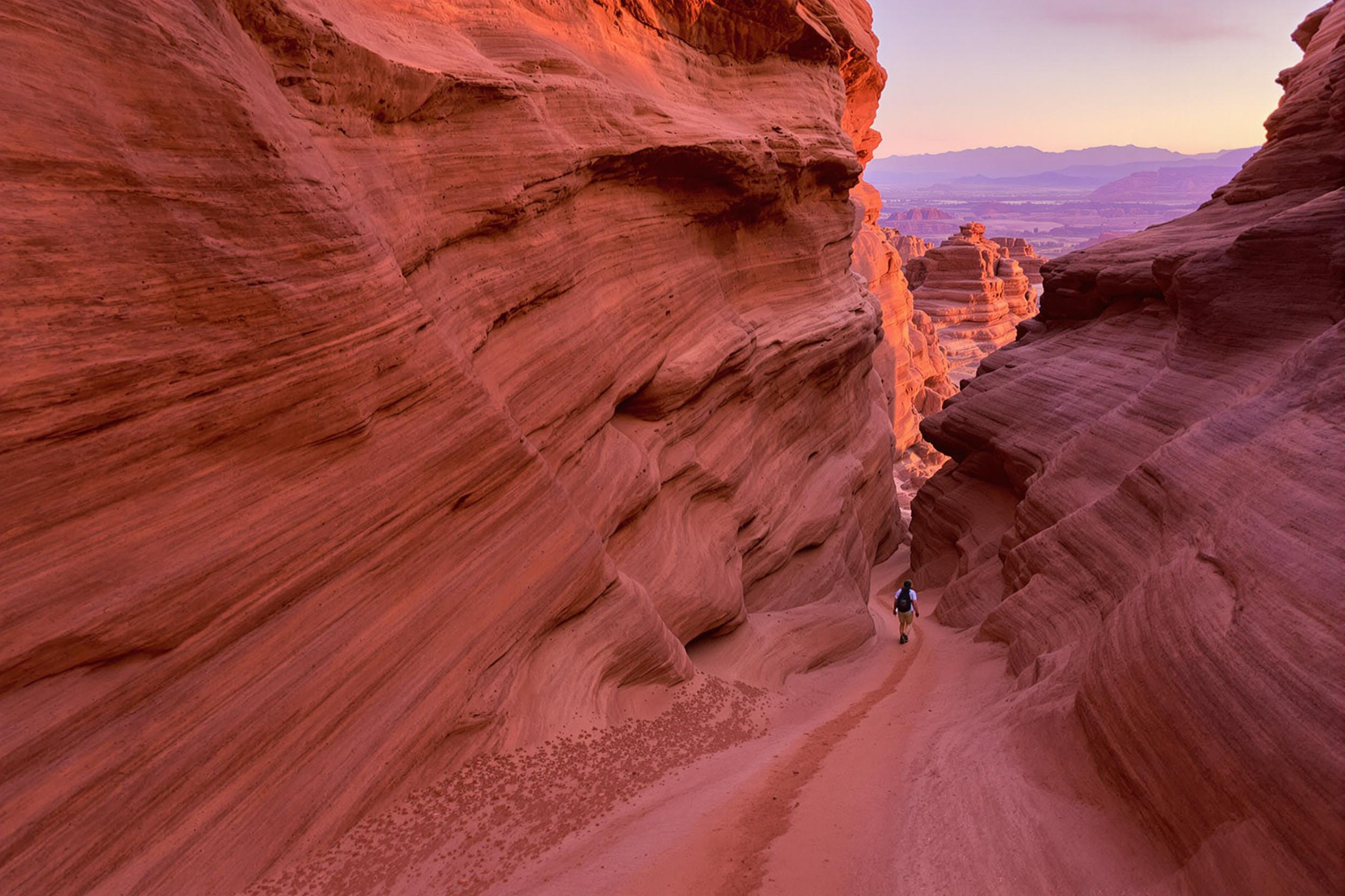 Amid the steep sandstone walls of a winding canyon, a lone traveler in desert attire strides into the fading dusk. Diffuse light enhances the warm orange and soft purple hues of the landscape, while faint silhouettes of distant ridgelines create a tranquil sense of scale. Wisps of fine sand trail in their wake, captured against smooth rock textures.