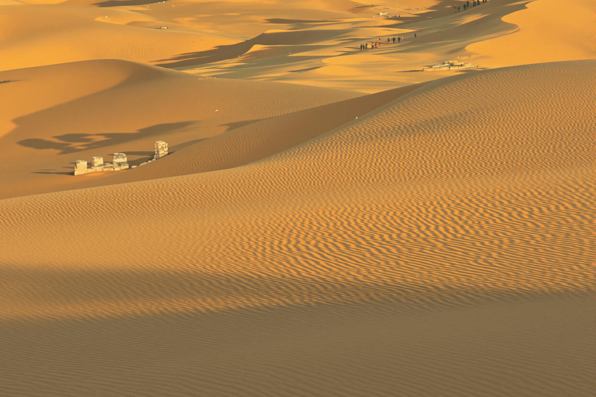 In a quiet desert expanse, ancient stone ruins rise above undulating dunes of golden sand. The remnants of weathered columns cast long shadows across the terrain, illuminated by warm, low-angle sunlight. The stark contrast between pale stone textures and soft, rippled sand evokes timeless solitude.