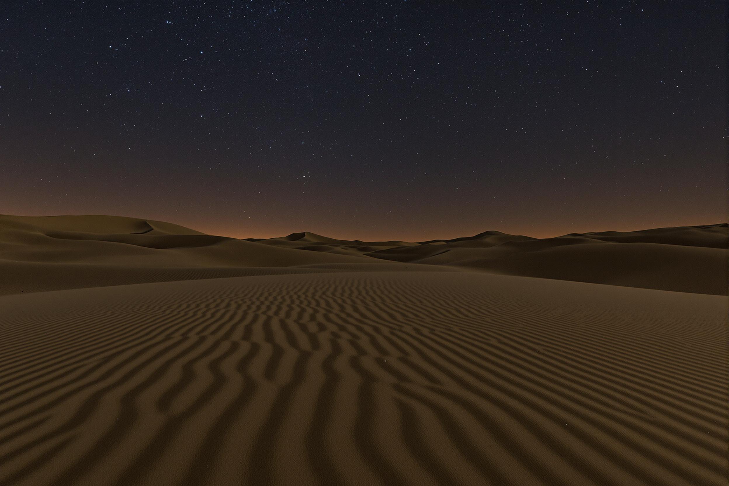 Under a clear night sky, vast desert dunes stretch out, their fine ripples accentuated by faint moonlight. The intricate textures of the sand contrast sharply against the dark sky adorned with countless shimmering stars. Subtle contours and flowing shapes of the landscape enhance the serene, infinite atmosphere.