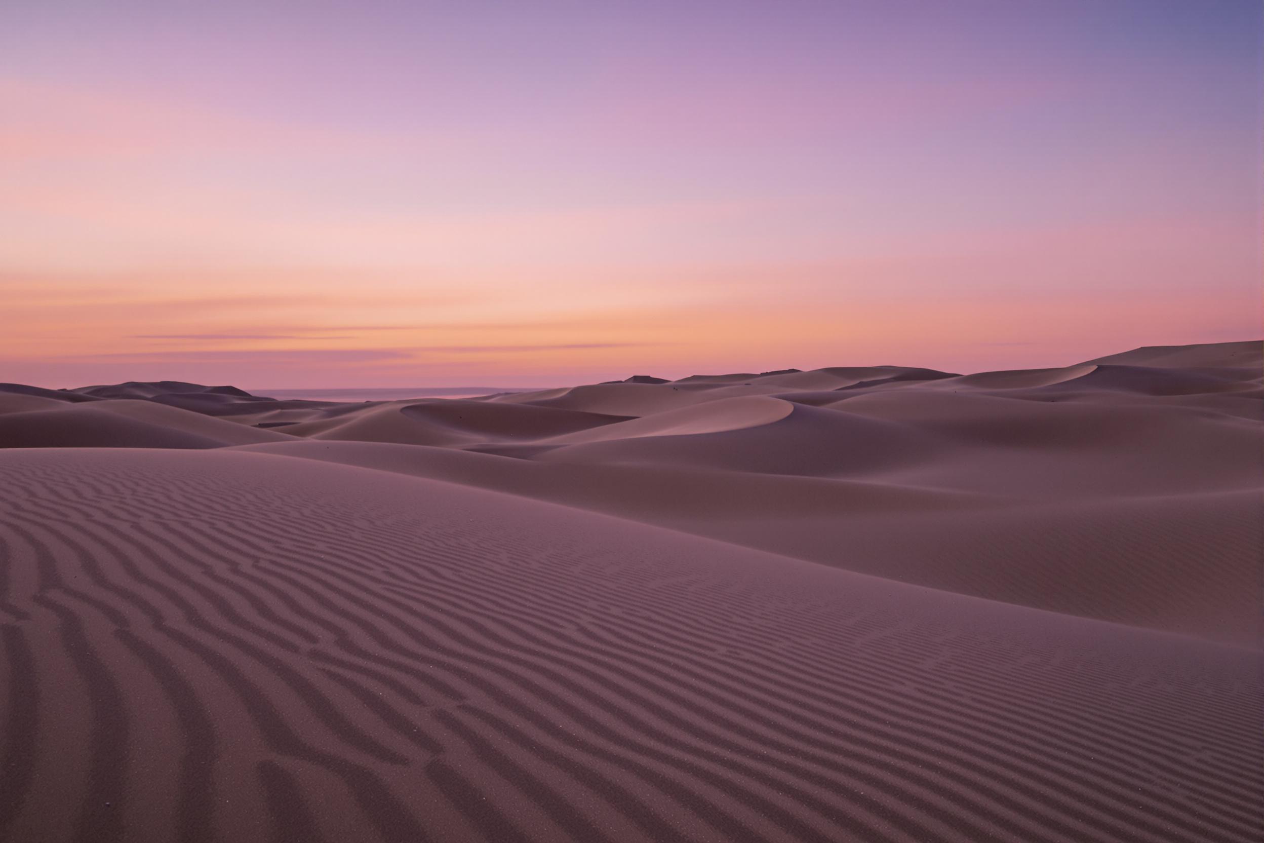 A tranquil scene captures desert sand dunes under a twilight sky. Soft pastels of purples and oranges paint the horizon, while gentle curves of sun-swept sand create flowing textures. Distant silhouetted shapes add depth to an otherwise minimalist and serene landscape, bathed in fading daylight.