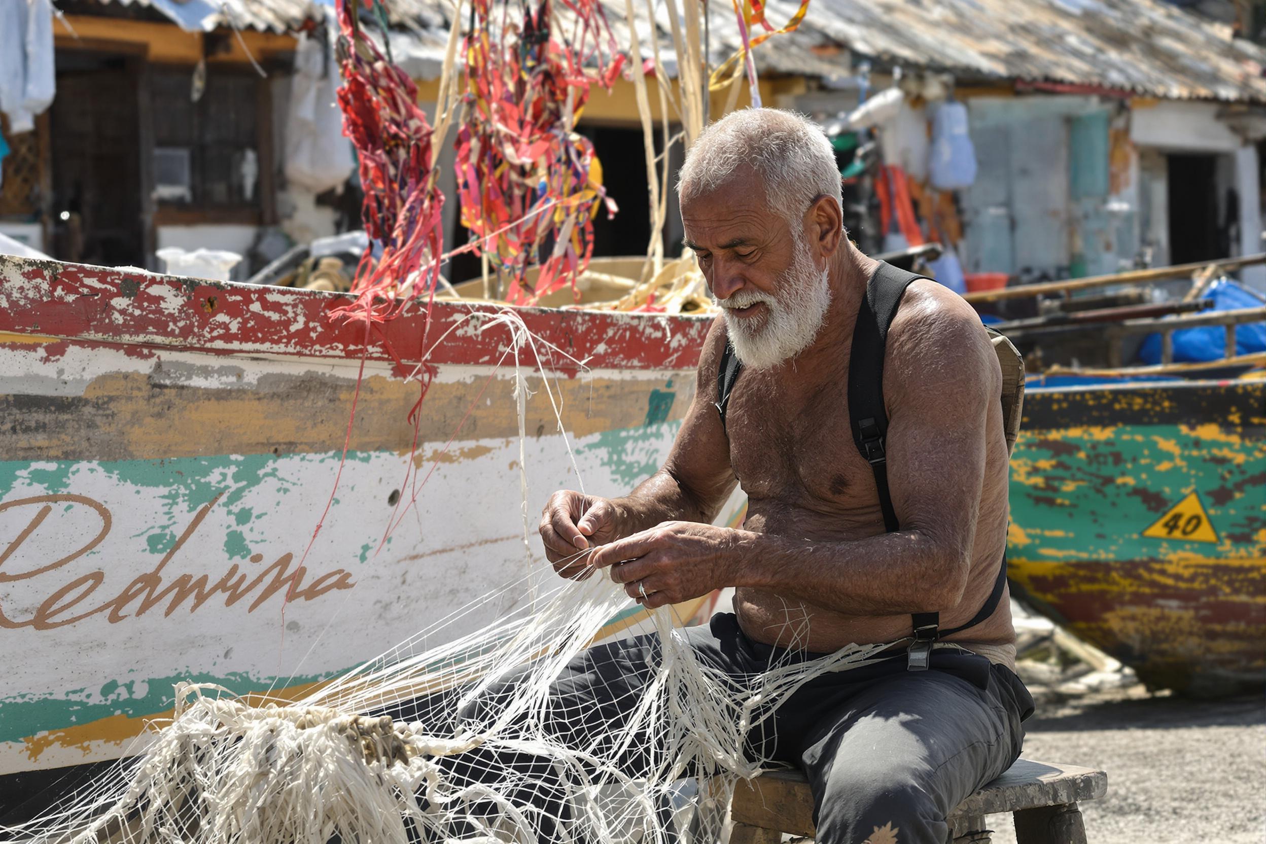 An elderly fisherman sits on a worn wooden stool in a sunlit coastal village. His wrinkled hands meticulously weave threads into a fishing net as sunlight catches the faded paint of his nearby boat. The backdrop features blurred rustic huts and fishing gear, adding depth and texture to the serene scene.