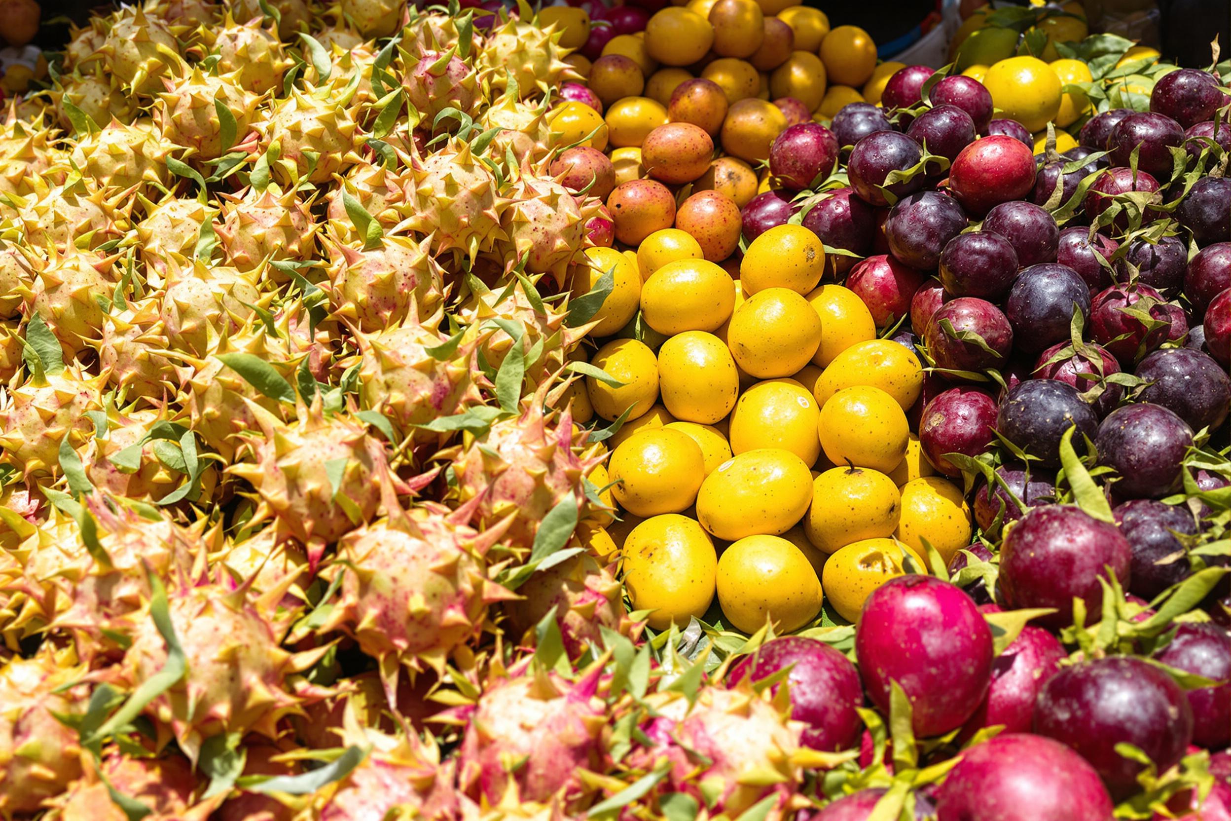 A close-up captures a vibrant array of exotic fruits at a bustling market stall. Lush dragon fruits, bright yellow mangosteens, and deep purple star fruits are arranged beautifully, showcasing their unique textures and intricate patterns. Brilliant sunlight illuminates the scene, emphasizing the freshness and rich colors that attract eager shoppers.