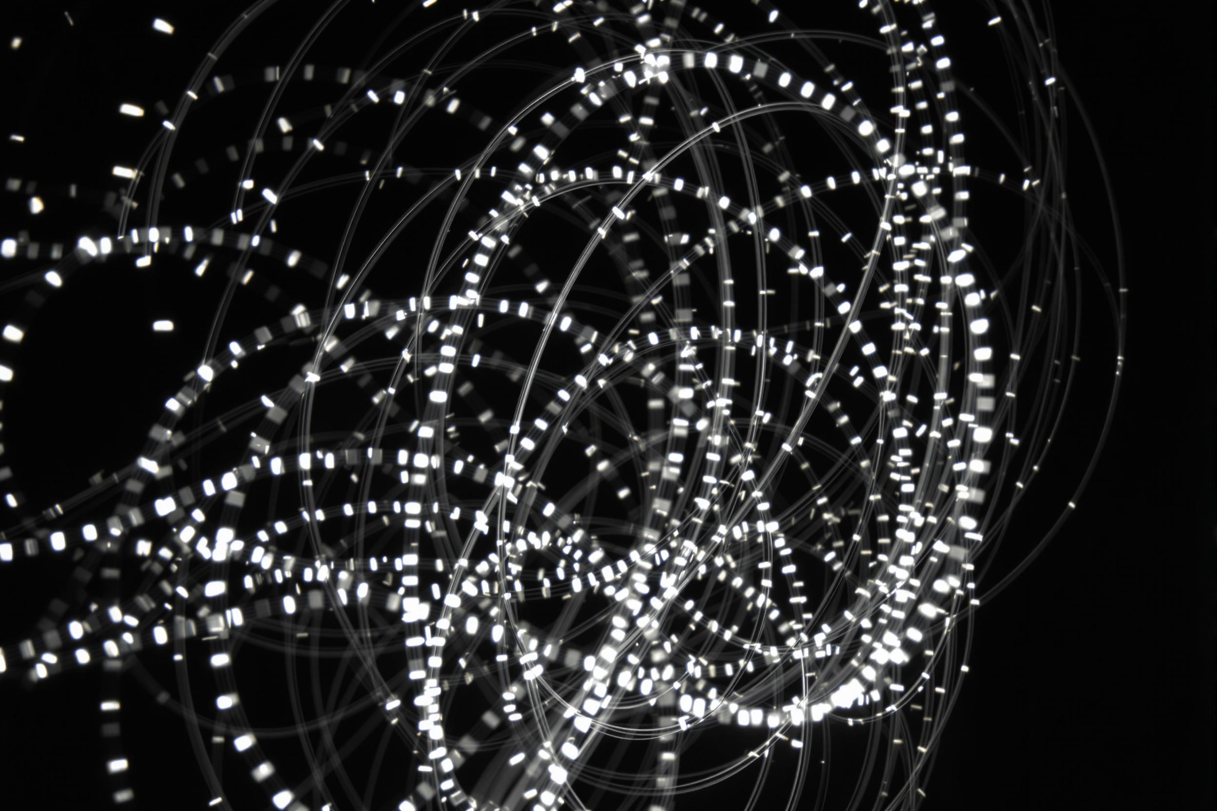 Capture the mesmerizing interplay of light and motion in this experimental photograph of a kinetic light sculpture. Graceful arcs of LED lights trace intricate patterns against a dark background, creating a hypnotic visual symphony of movement and illumination.