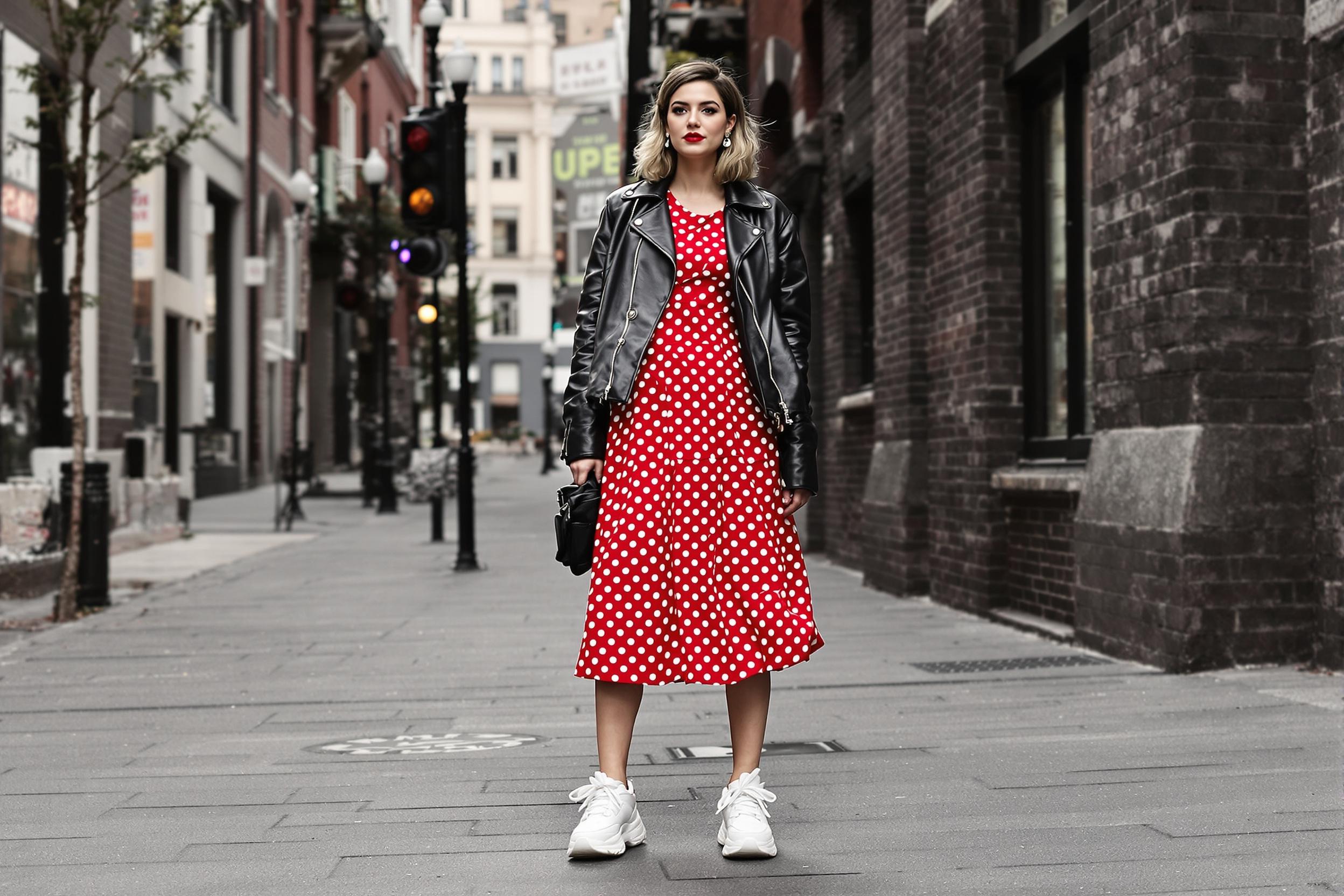 A striking fusion of eras captured in an urban landscape. A model sports a 1950s-inspired polka dot dress, artfully paired with a contemporary leather jacket and chunky sneakers. The juxtaposition of vintage charm and modern edge creates a captivating street style narrative.