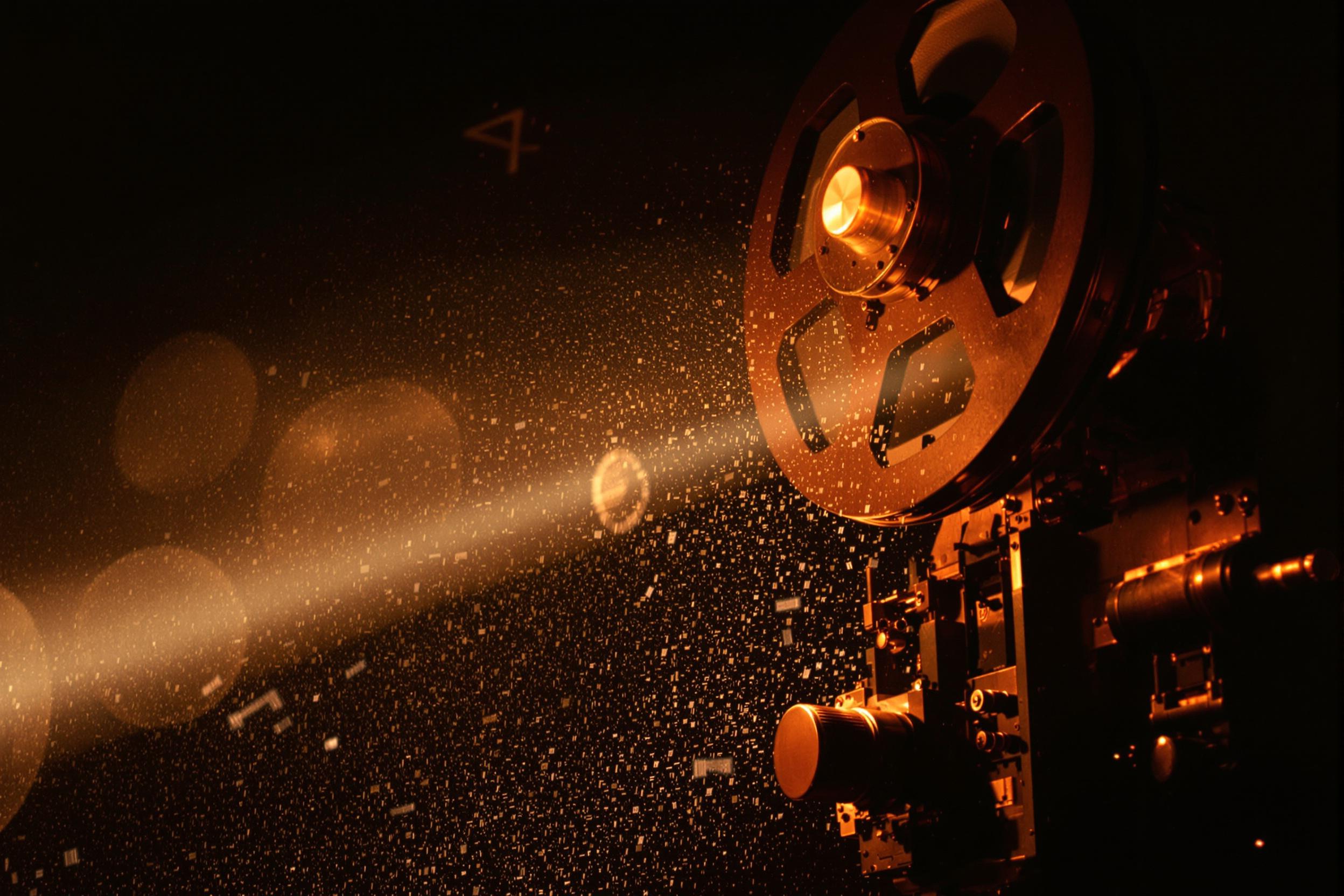 A classic 35mm film projector illuminates a dark room, casting a warm glow. The projector's mechanical parts are in motion, threading celluloid through its reels. Dust particles dance in the beam of light, creating a nostalgic atmosphere.