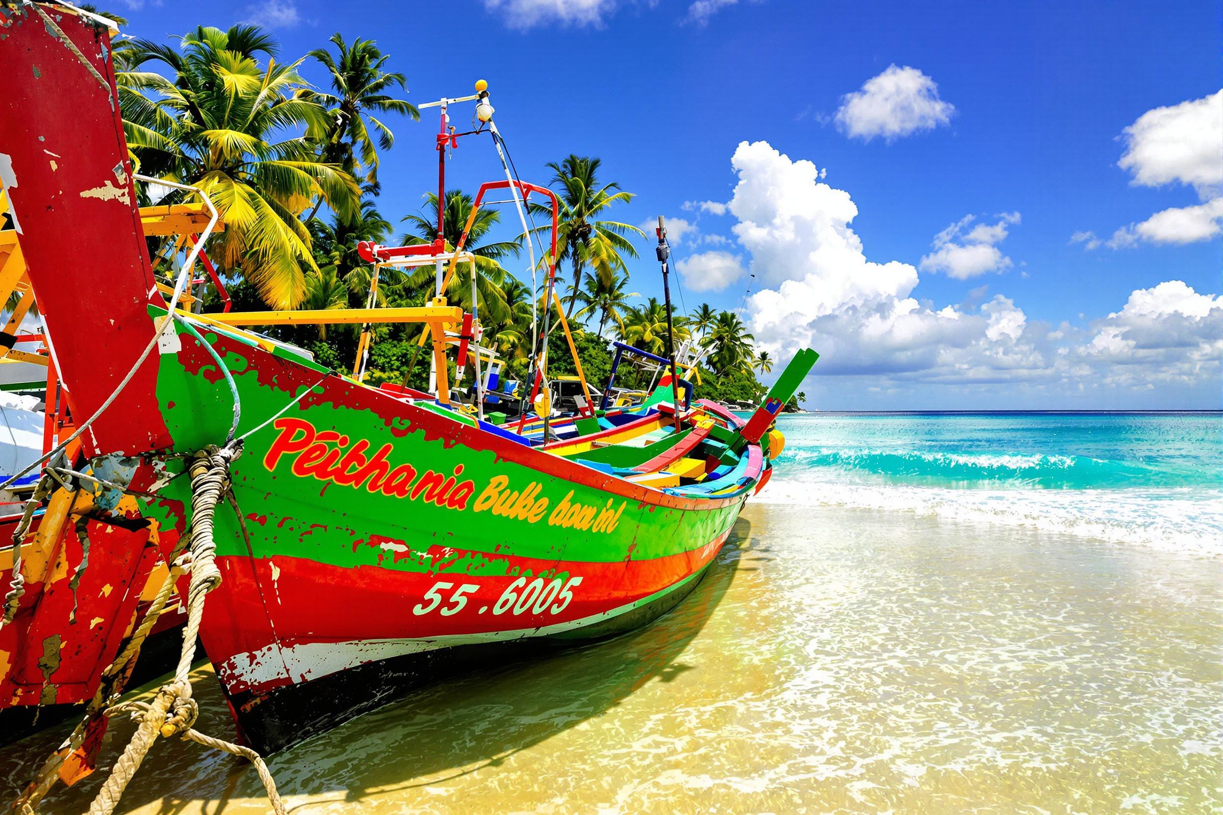 Vibrant fishing boats painted in bright hues of red, green, and yellow rest docked along a tropical shoreline. Turquoise waves gently lap against their worn hulls, reflecting golden sunlight. Distant palm trees frame the horizon under a cloudless azure sky, while soft sand stretches along the tranquil scene.